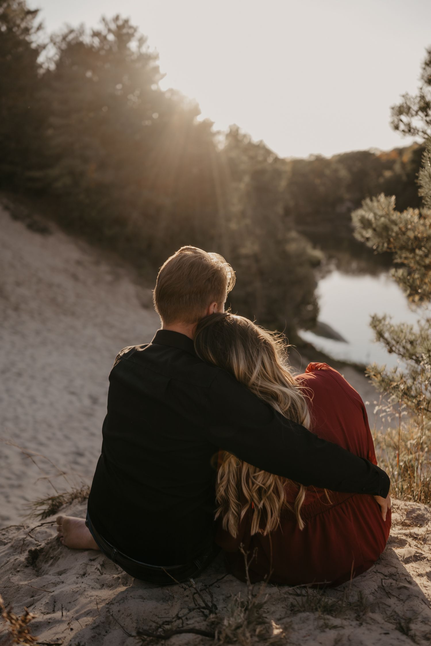 Couple sitting together on sand dunes watching golden sunset over river surrounded by pine trees.