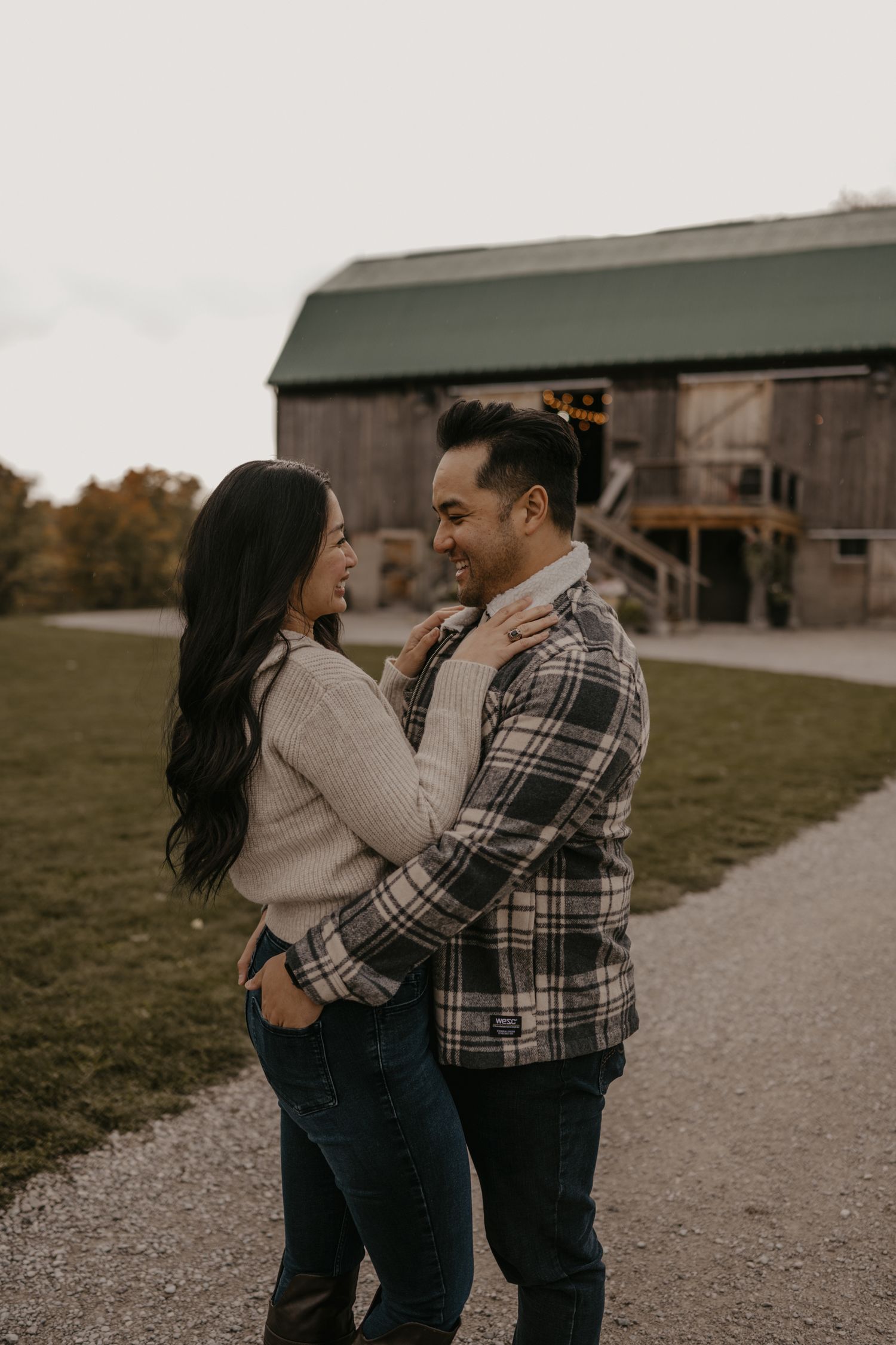 Couple embracing and smiling at each other outdoors near a rustic barn in autumn, wearing cozy flannel and knit sweater.
