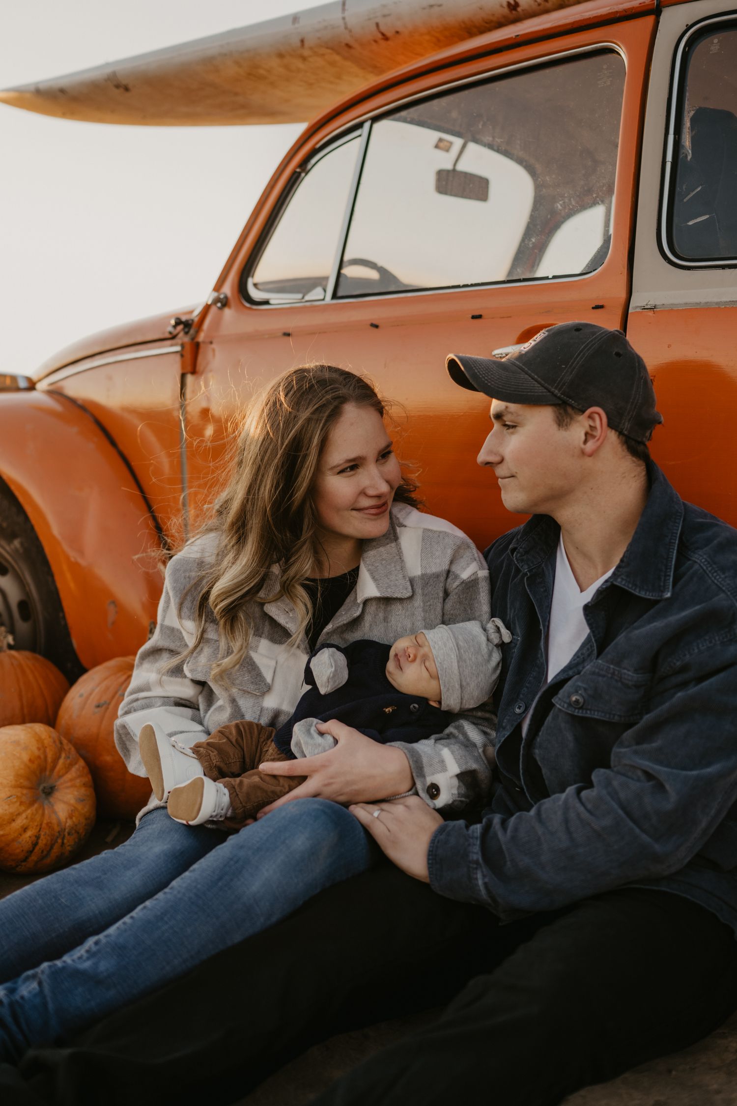 Young couple with newborn baby sitting against a rustic orange car surrounded by pumpkins.