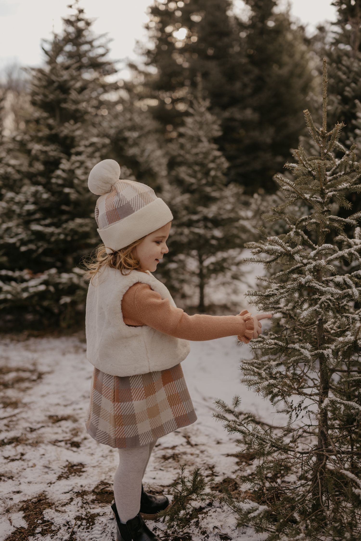 Young girl in plaid skirt and pom-pom hat touching a snow-dusted pine tree at a Christmas tree farm.
