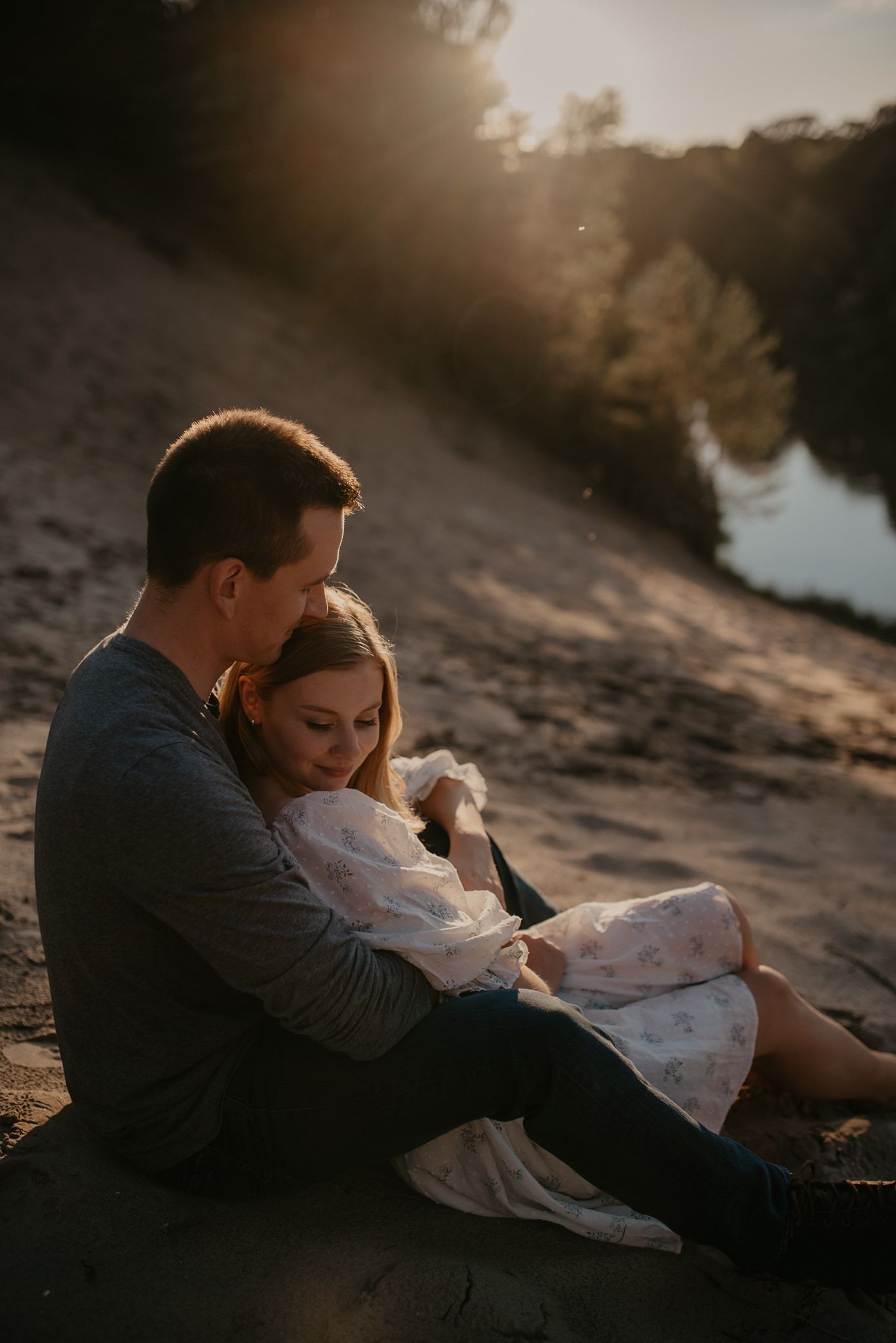 Couple sitting together on sandy riverbank at golden sunset, man embracing woman tenderly.