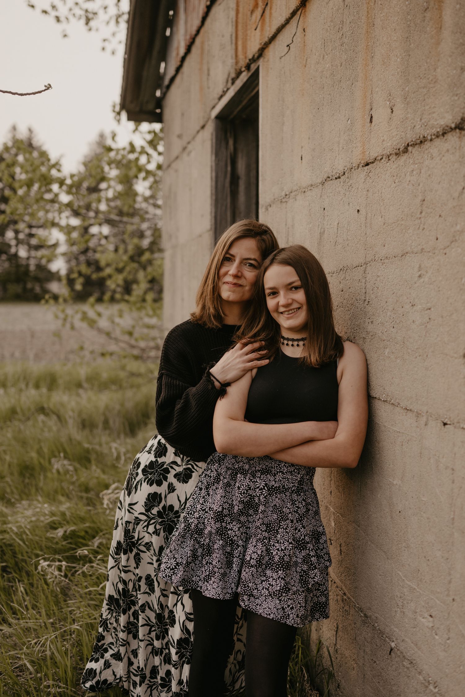 Two women pose against a rustic concrete wall outdoors, wearing floral skirts and black tops.