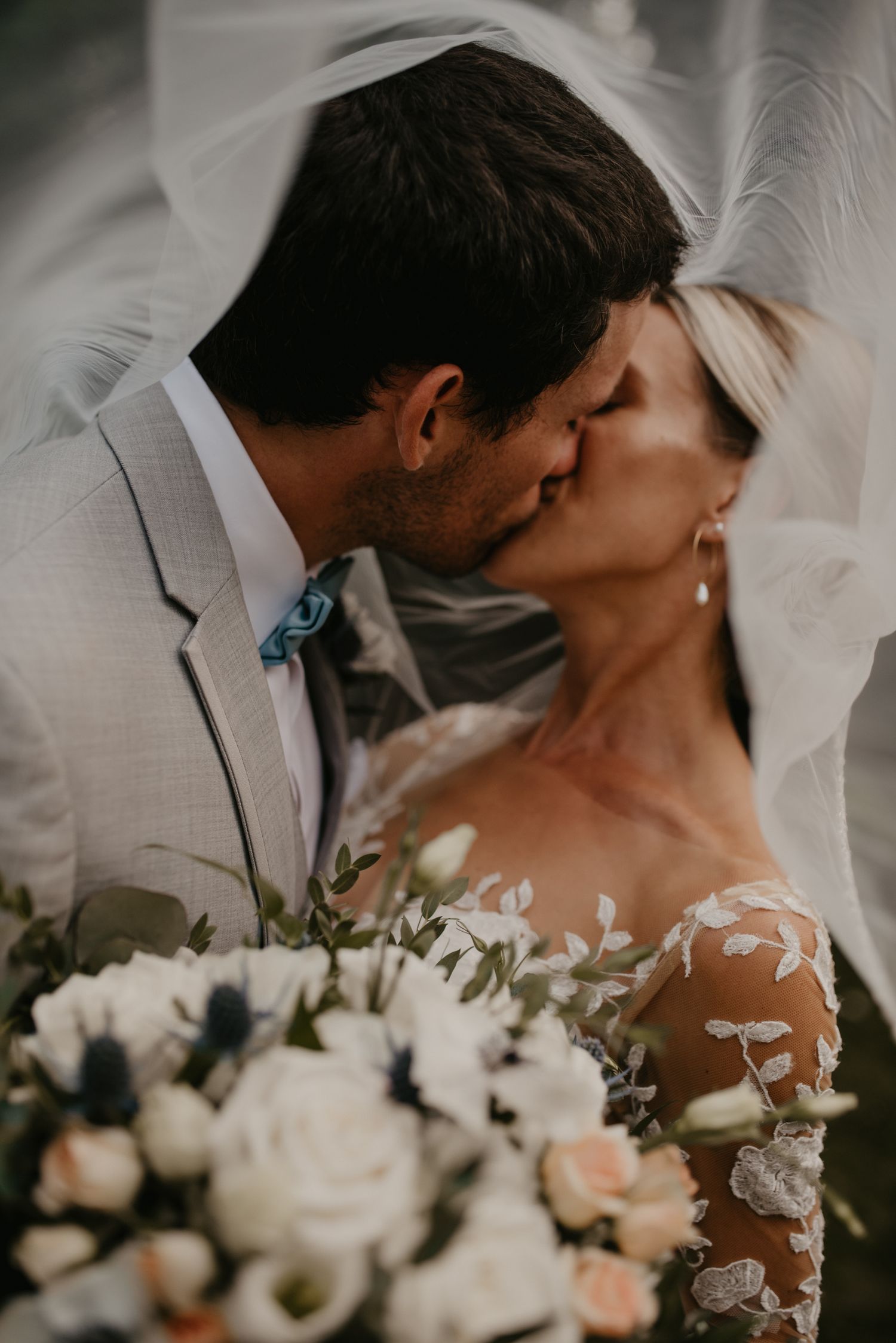 Bride and groom share a tender kiss beneath flowing veil with white floral bouquet.