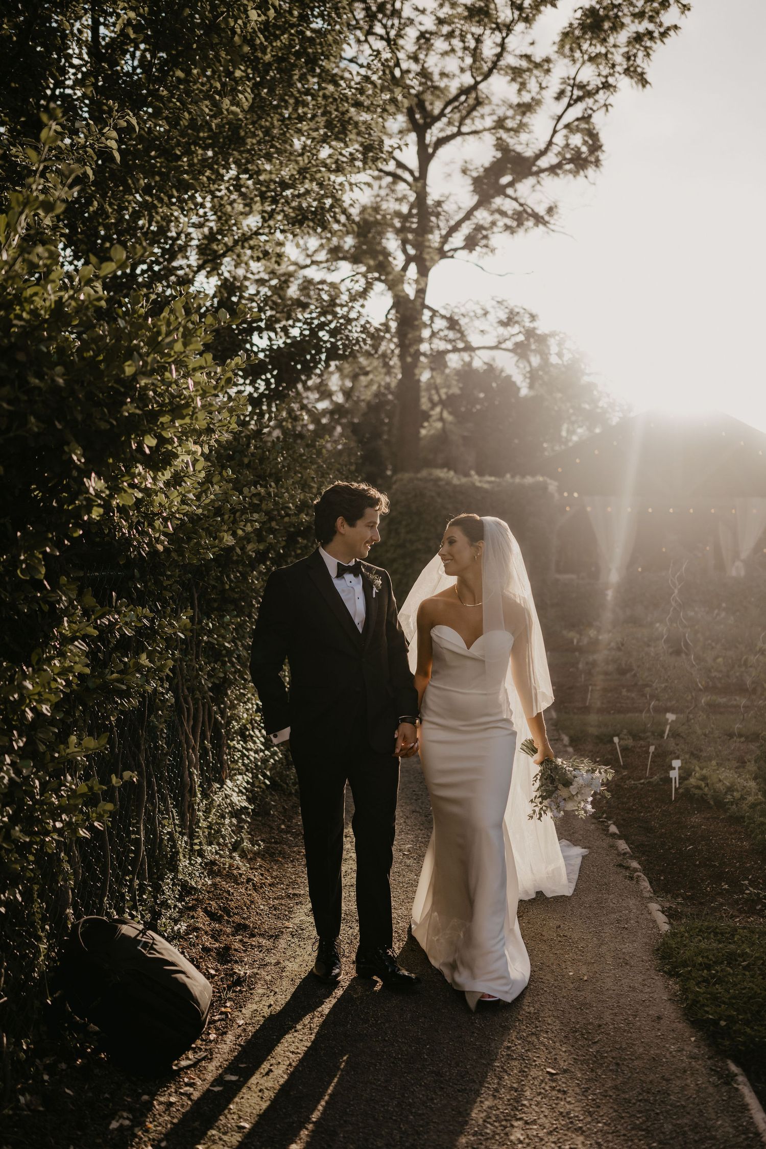 Newlyweds walk hand in hand along sunlit garden path in elegant wedding attire.