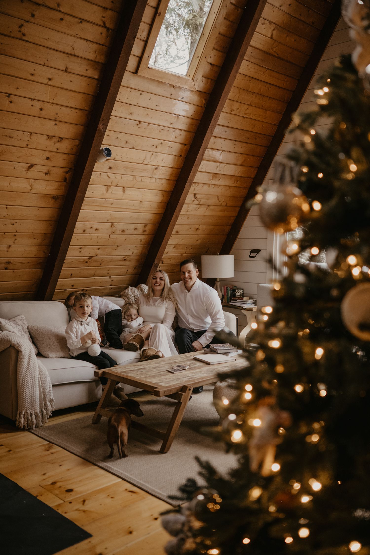 Family of four relaxing on a white sofa in a cozy A-frame cabin with wood walls and a decorated Christmas tree.