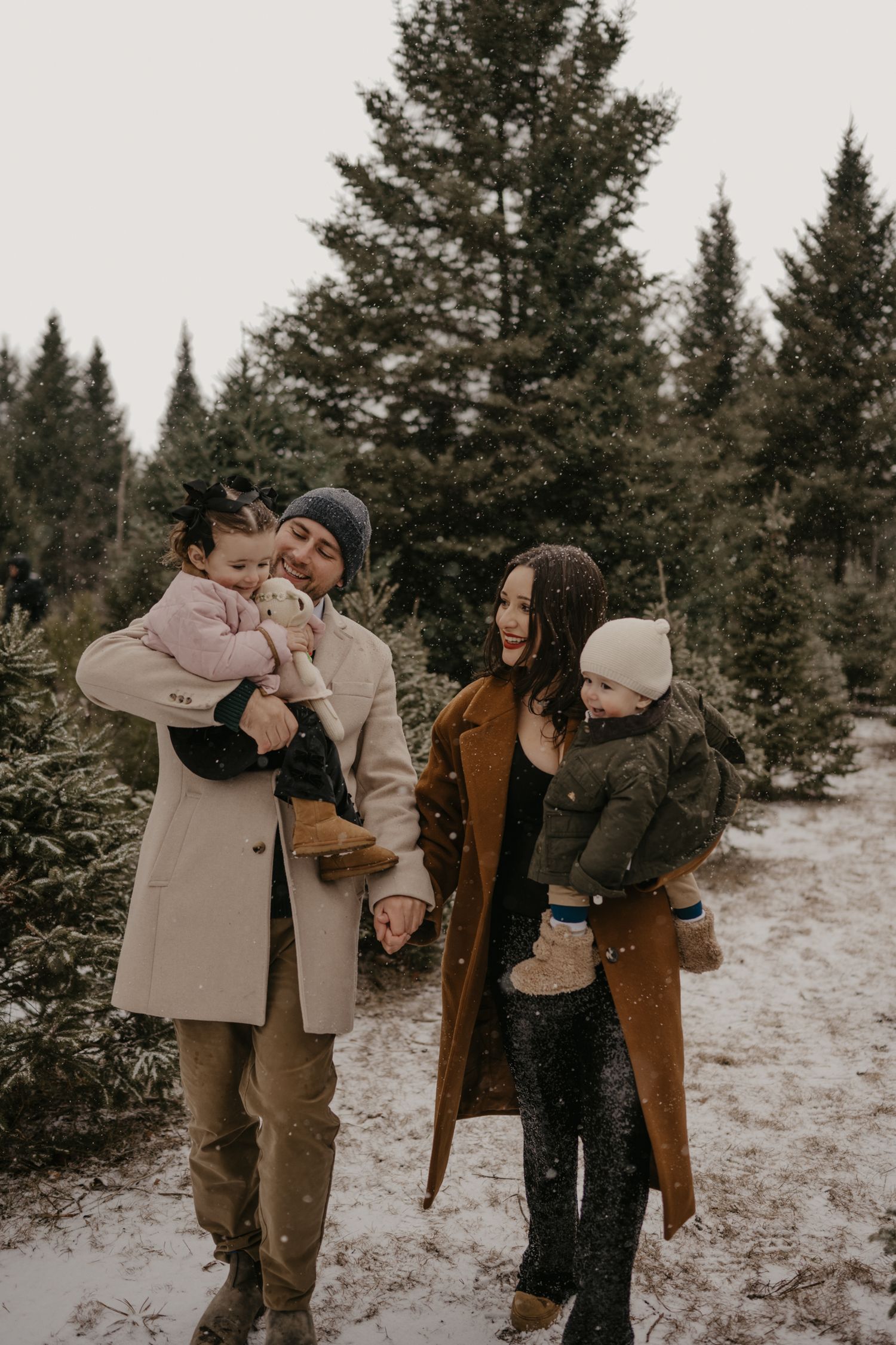 Family of four poses in snowy Christmas tree farm, parents holding toddlers in winter coats and beanies.