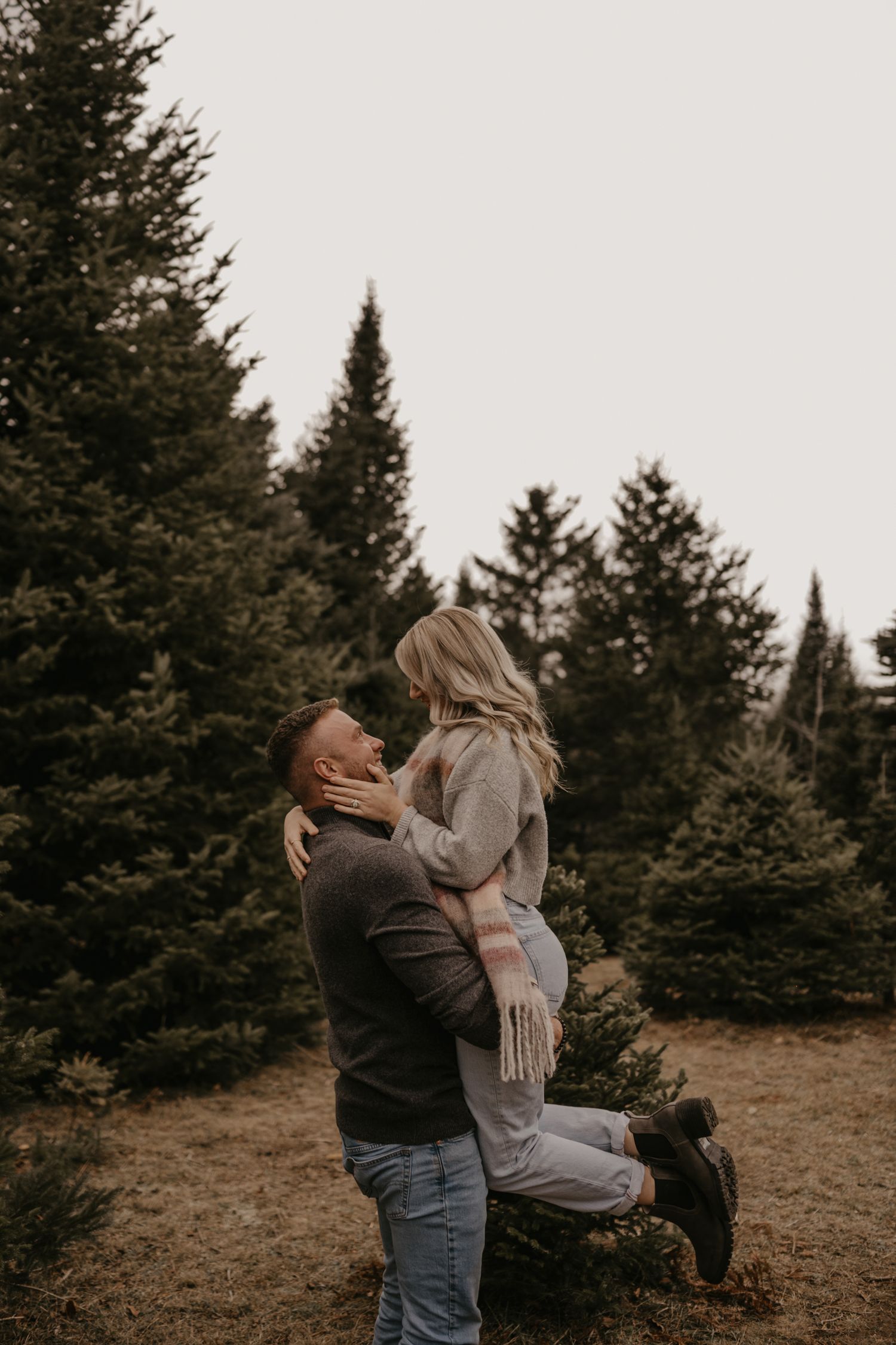 A man lifts a woman in a cozy plaid scarf among evergreen trees in a romantic outdoor couples photo.
