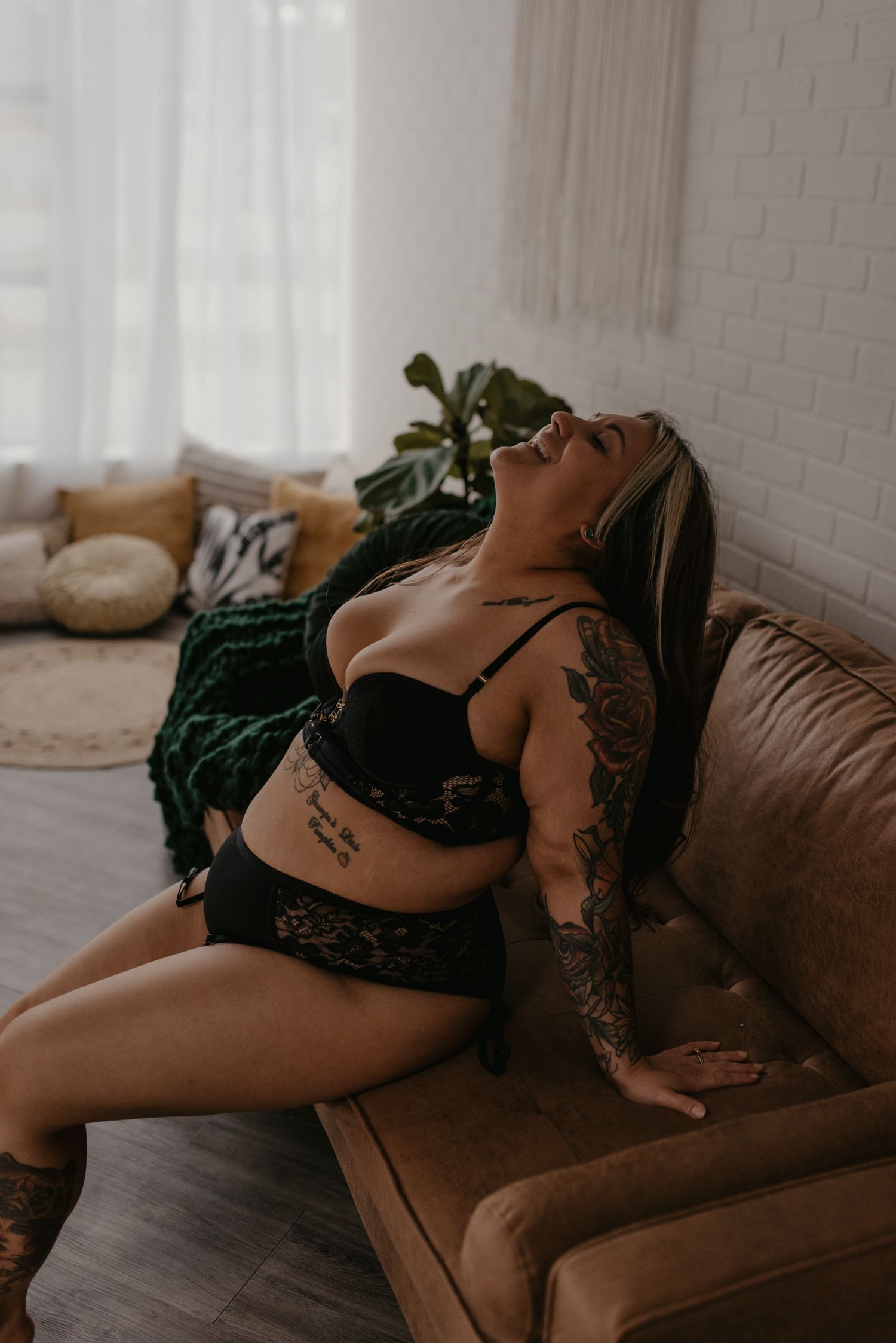 Tattooed blonde woman in black lingerie posing on leather couch in bright natural light boudoir shoot.