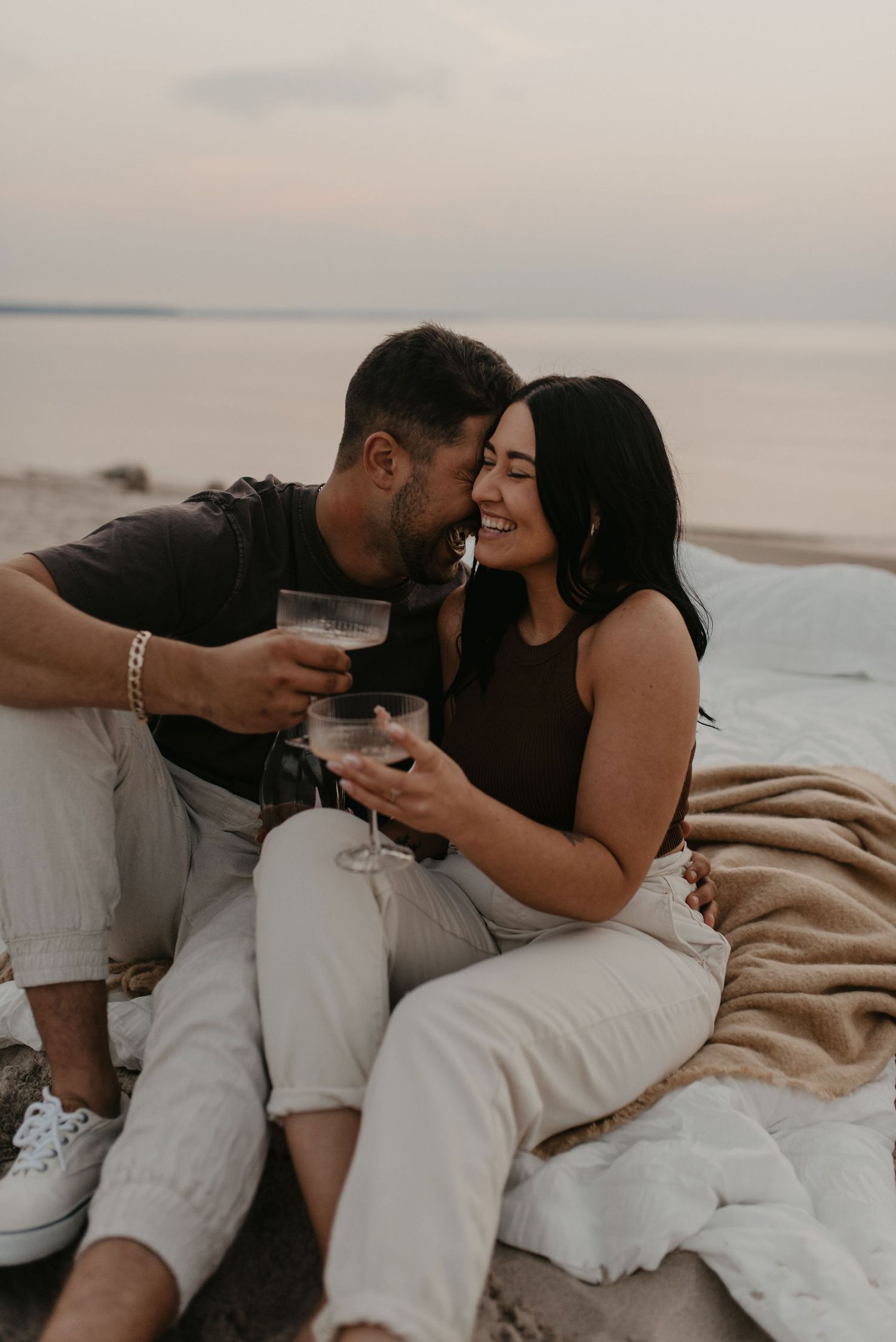 Romantic couple sharing wine glasses and kissing on beach blanket at sunset engagement session.