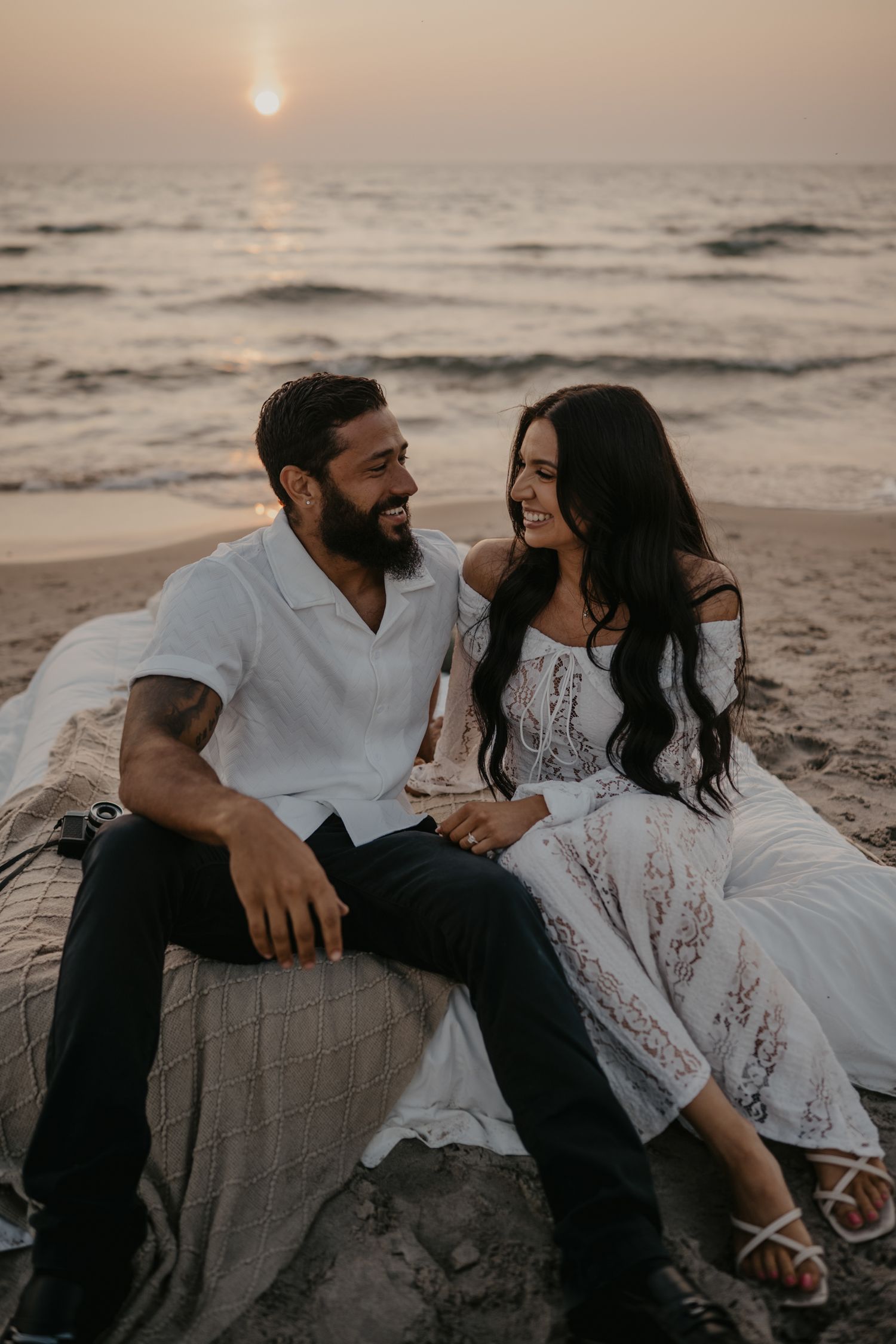 Couple sitting on beach blanket smiling at each other during a beautiful ocean sunset.