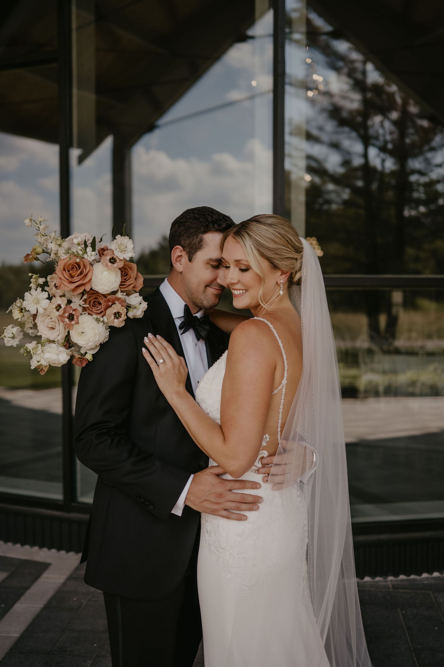 Bride and groom embracing outdoors, she in a white gown with veil, he in a black tuxedo, holding a floral bouquet.