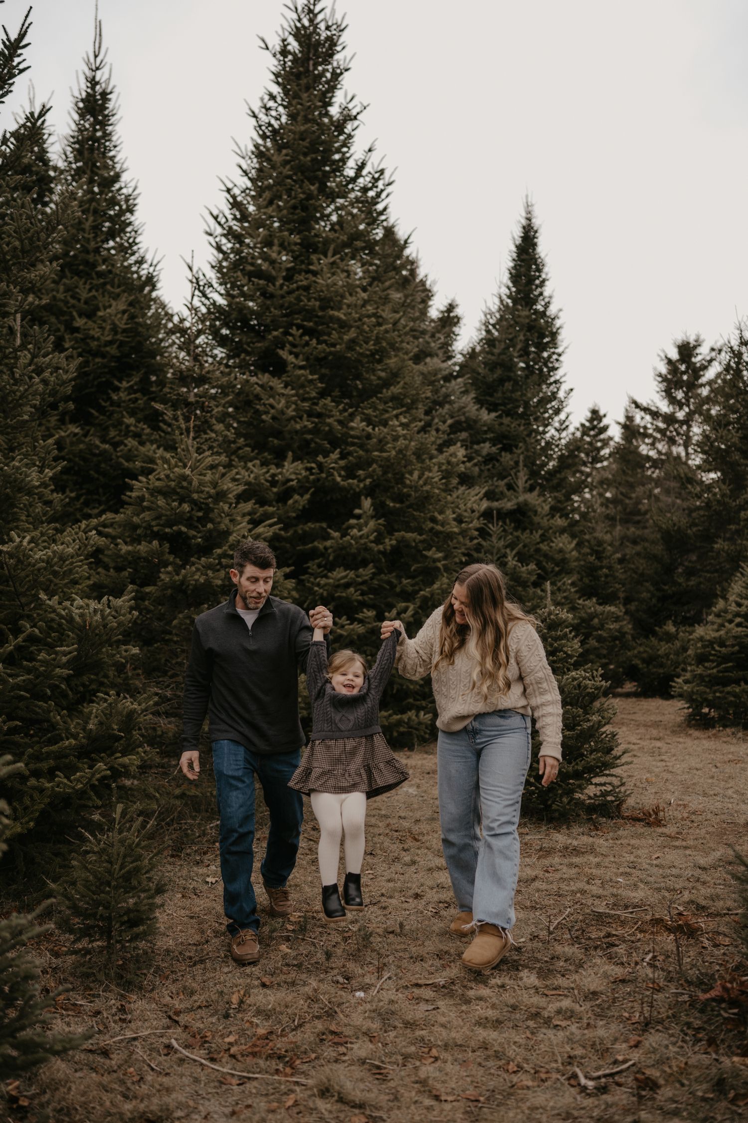 A family of three walks hand-in-hand through a Christmas tree farm, swinging their toddler between them.