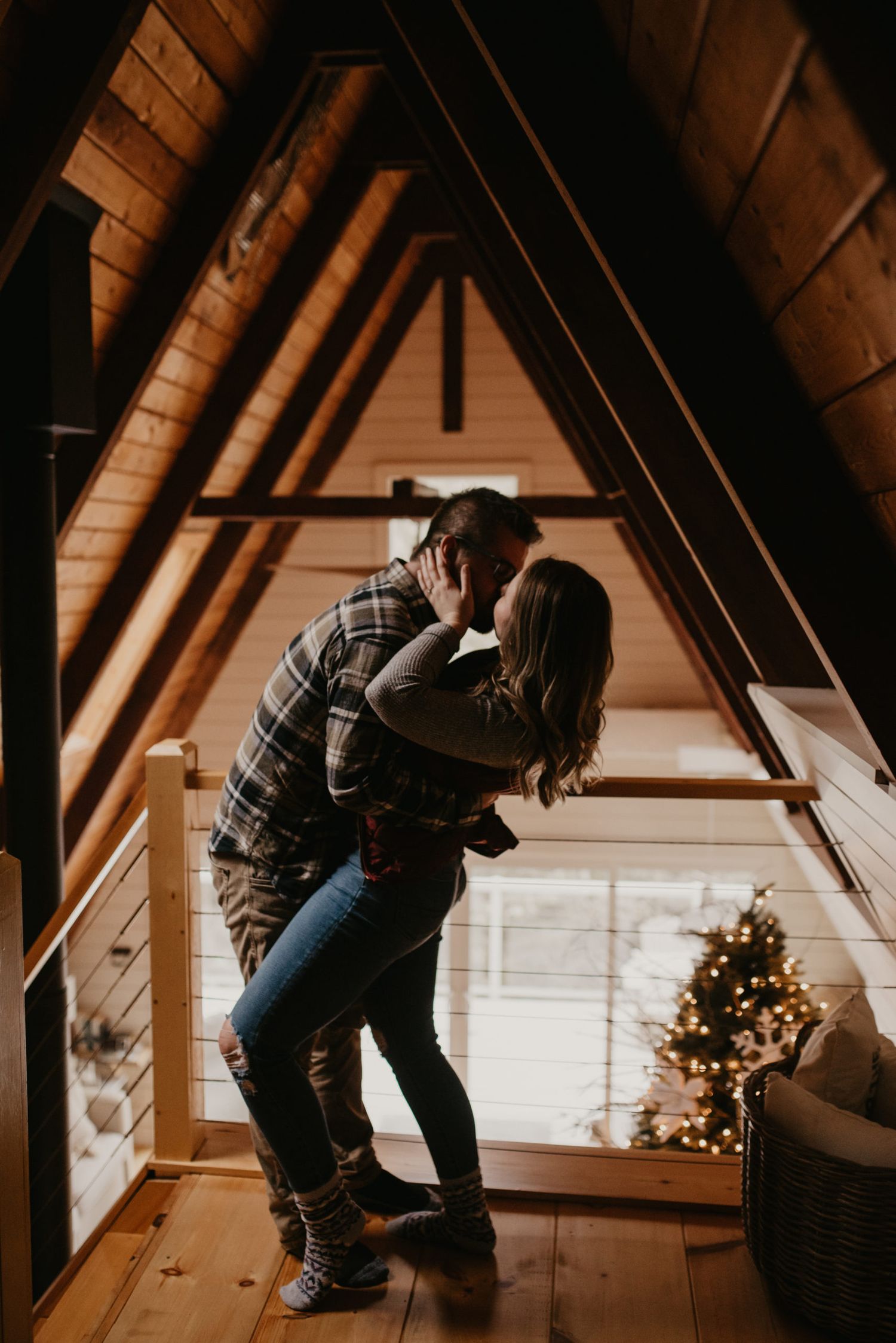 Couple sharing a romantic kiss in a cozy A-frame cabin loft with Christmas tree lights glowing in background.