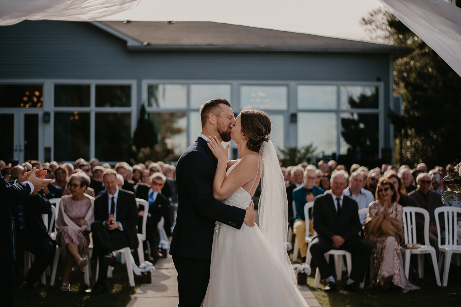 Bride and groom sharing first kiss during outdoor wedding ceremony with guests seated behind.