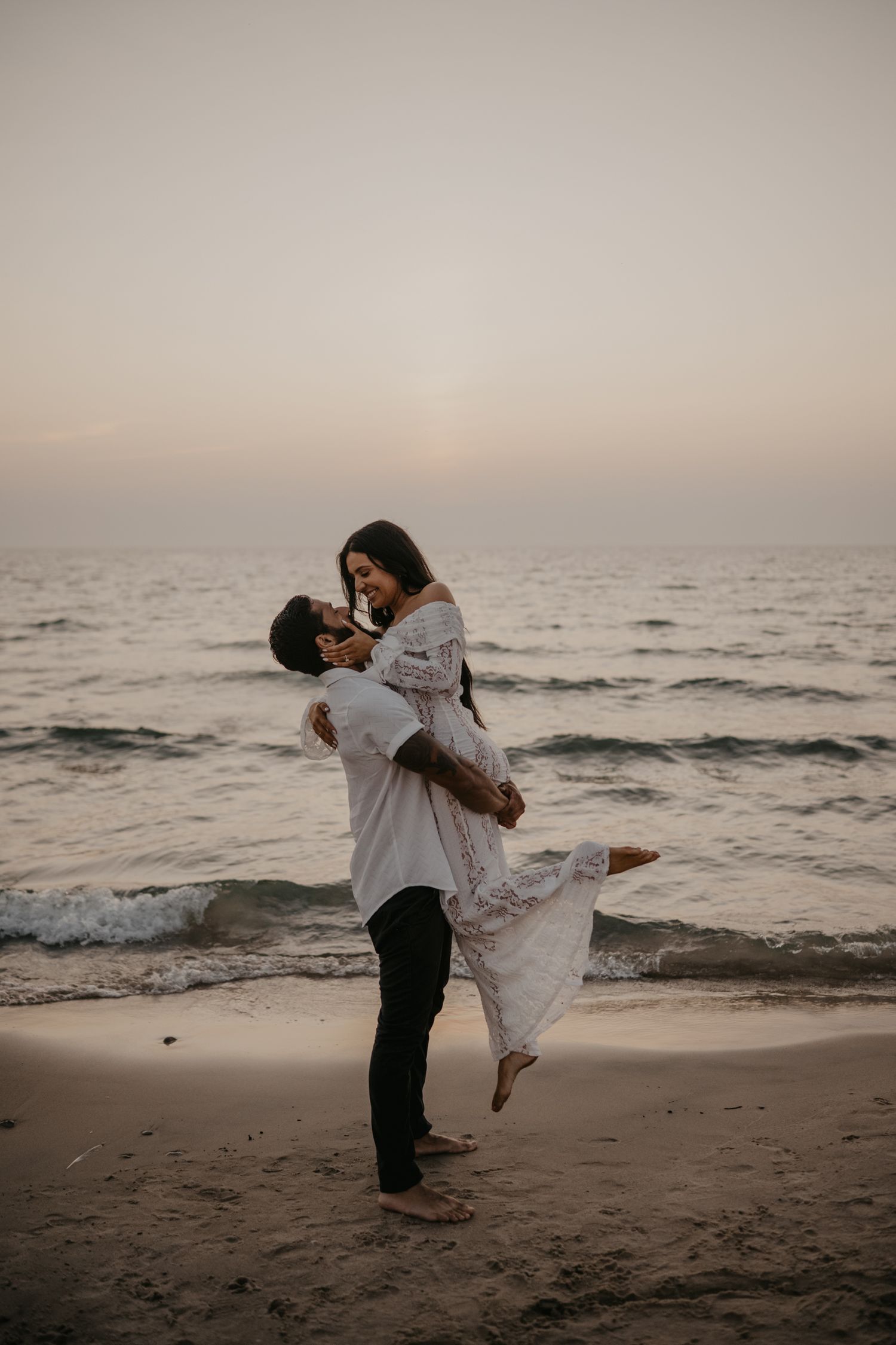 Man lifting woman in his arms on sandy beach at sunset by the ocean.