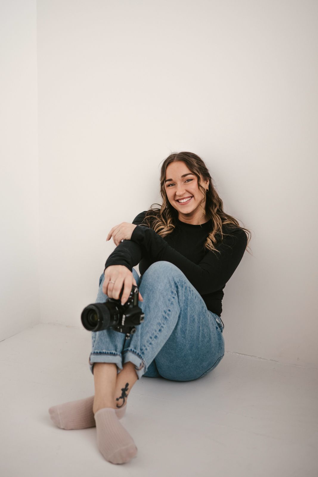 Smiling young woman sitting on floor holding a DSLR camera, wearing black sweater and jeans.