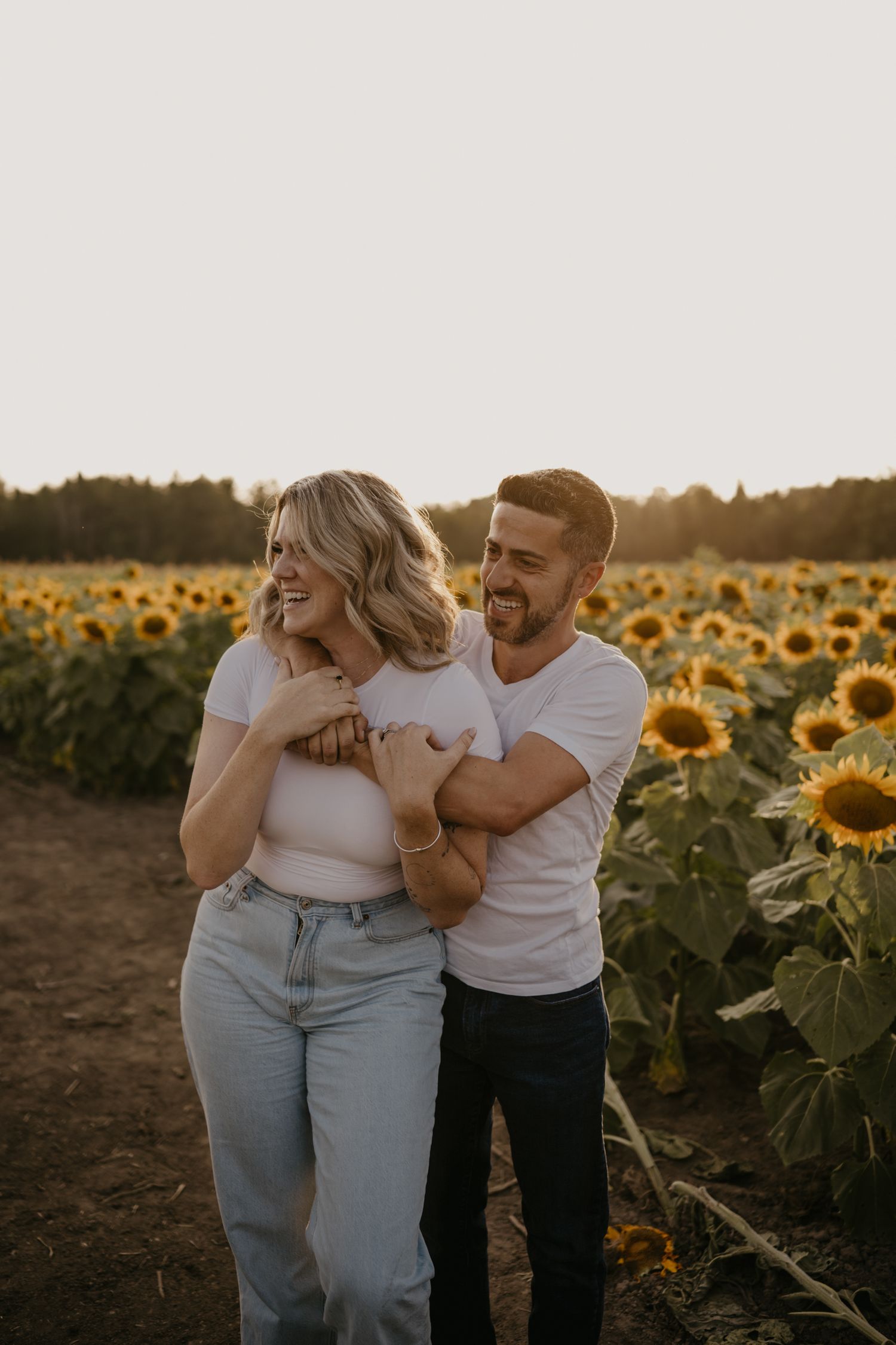 Man hugging woman from behind while laughing in a vibrant sunflower field at sunset.
