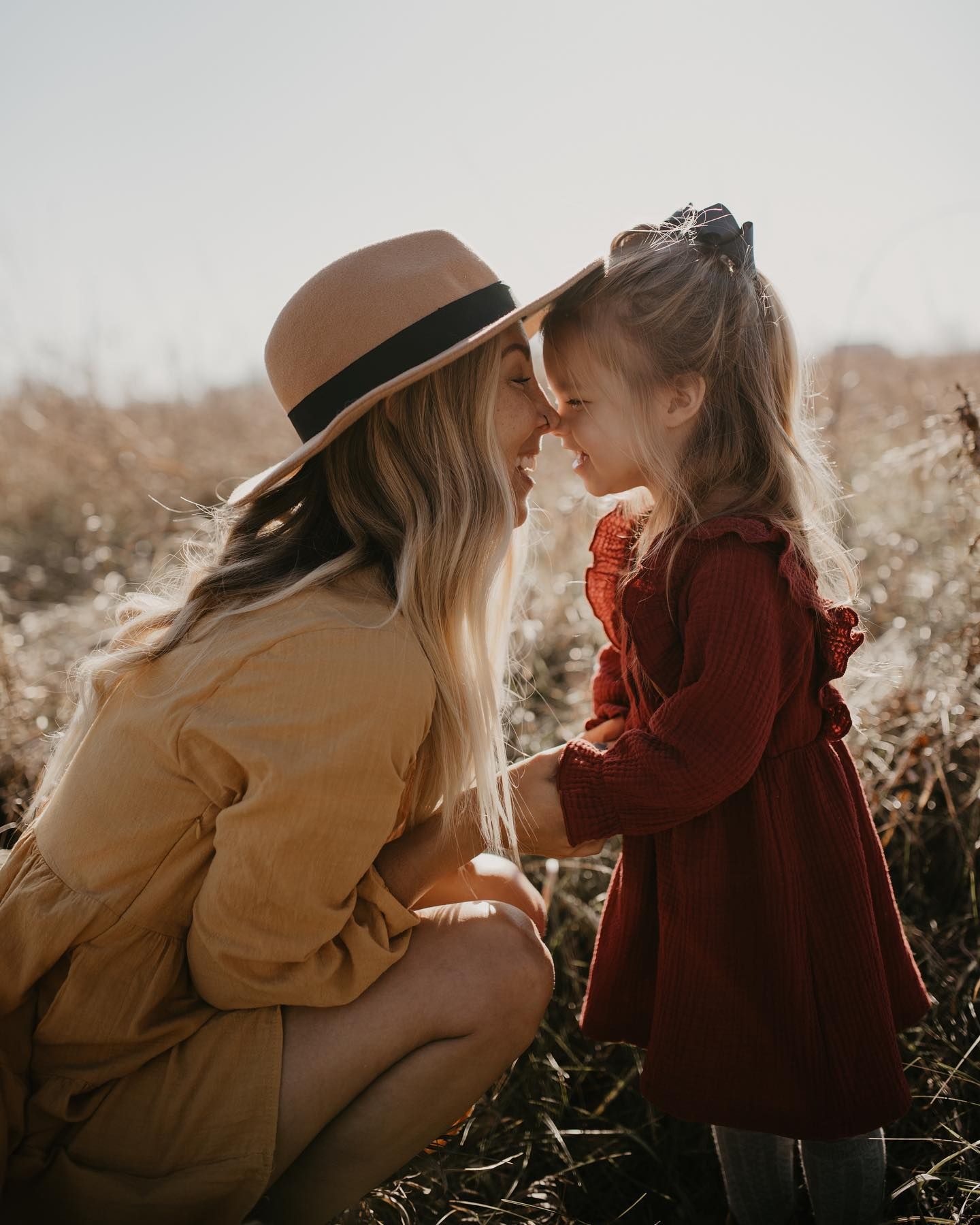 Mother in tan hat and dress touching noses with her young daughter in a red dress in a sunny field.
