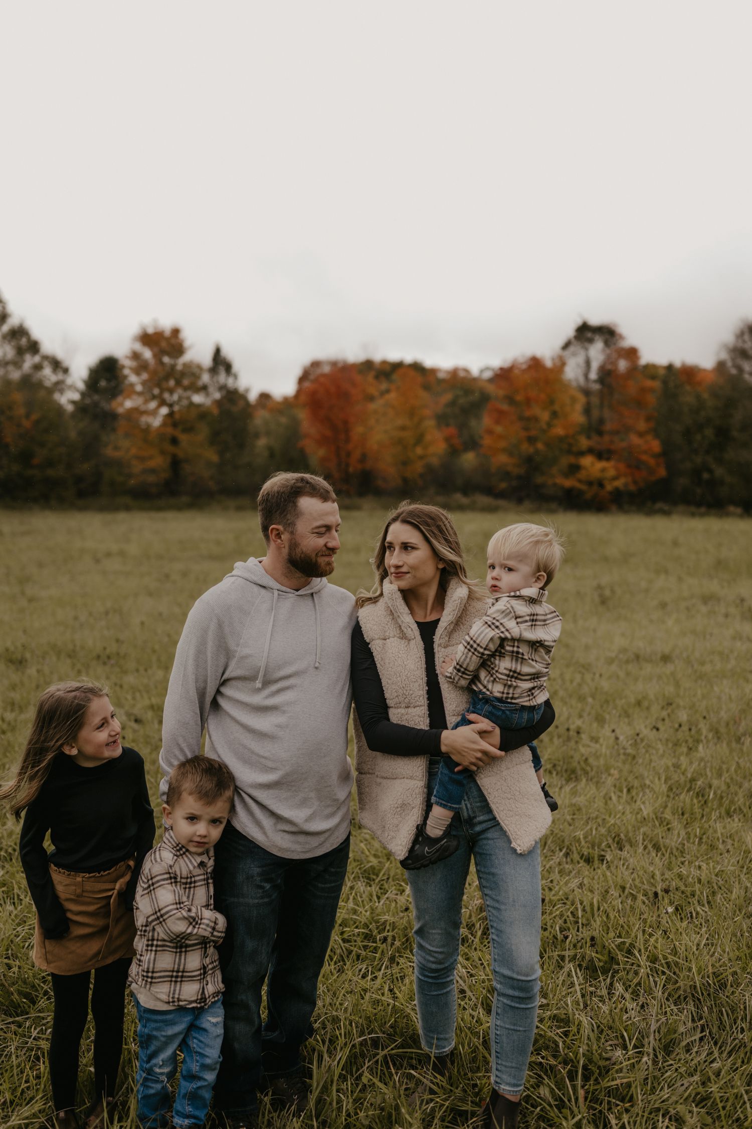 Family of five poses in autumn field with colorful fall foliage backdrop.