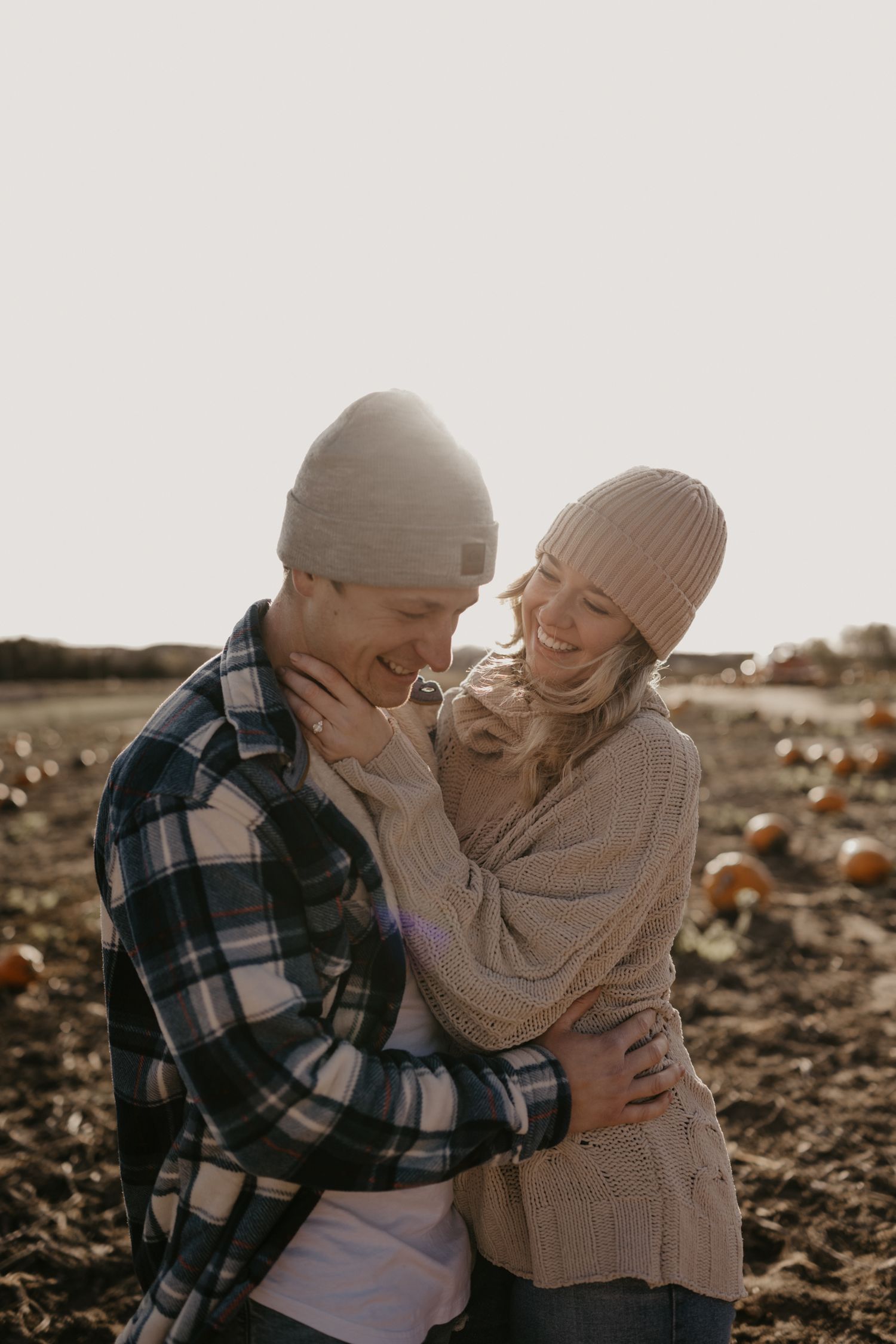 A couple laughs and embraces in a pumpkin patch, wearing cozy knit beanies and flannel in warm autumn sunlight.
