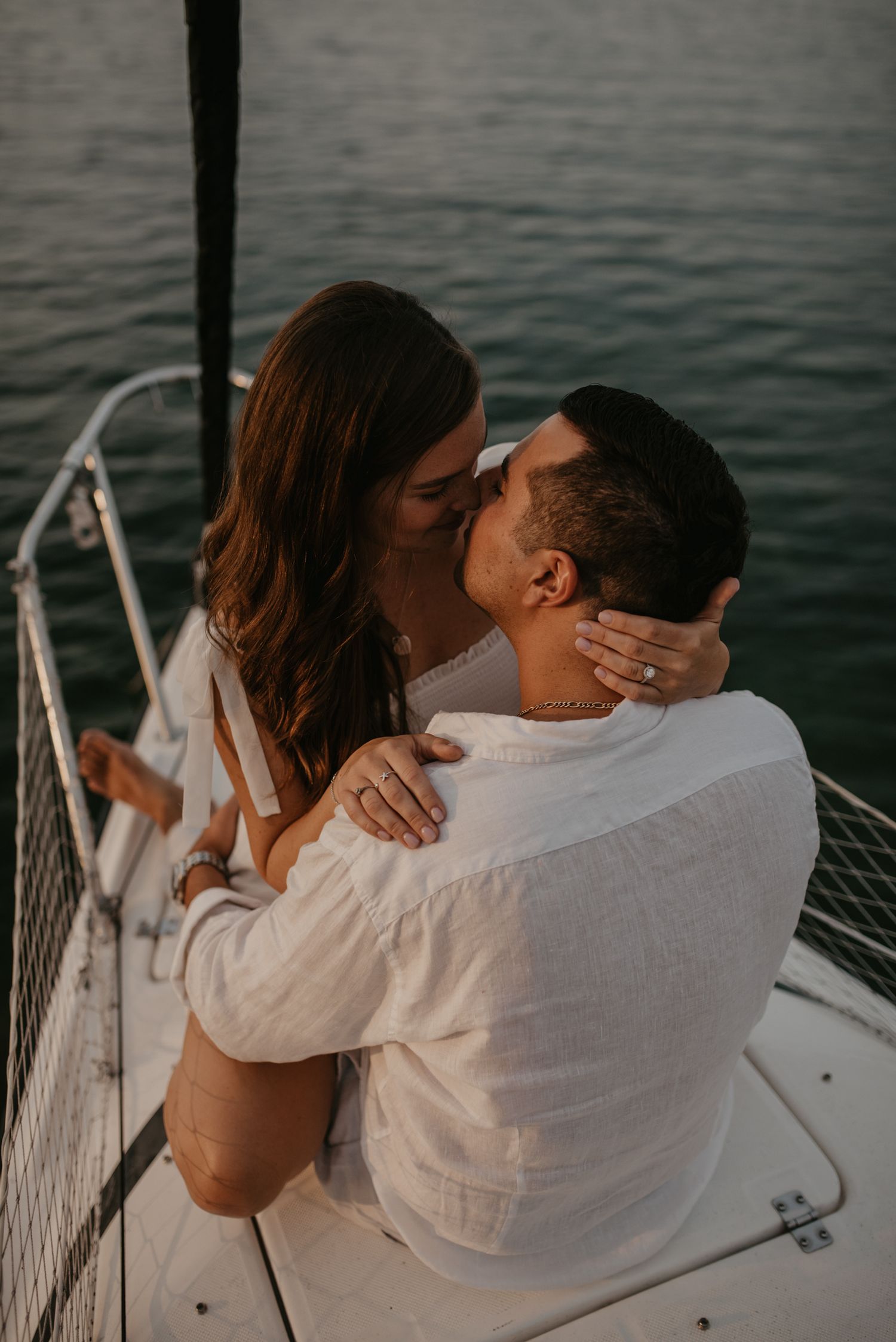 Couple kissing and embracing on a sailboat with calm green water in the background at sunset.