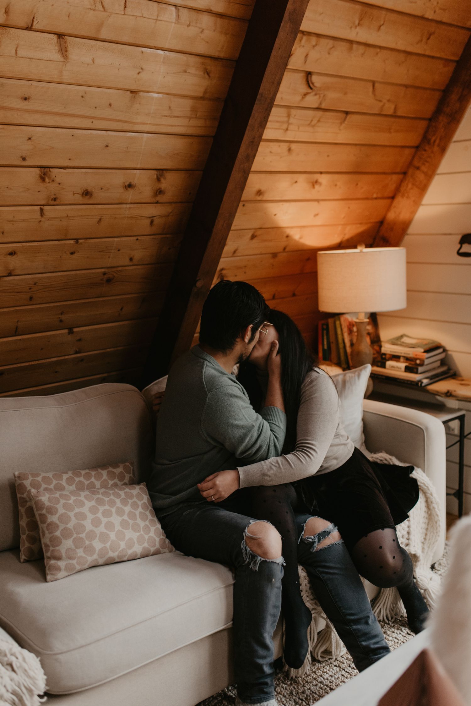 Couple sharing an intimate moment on a white sofa in a cozy wooden cabin loft with warm lamp light.