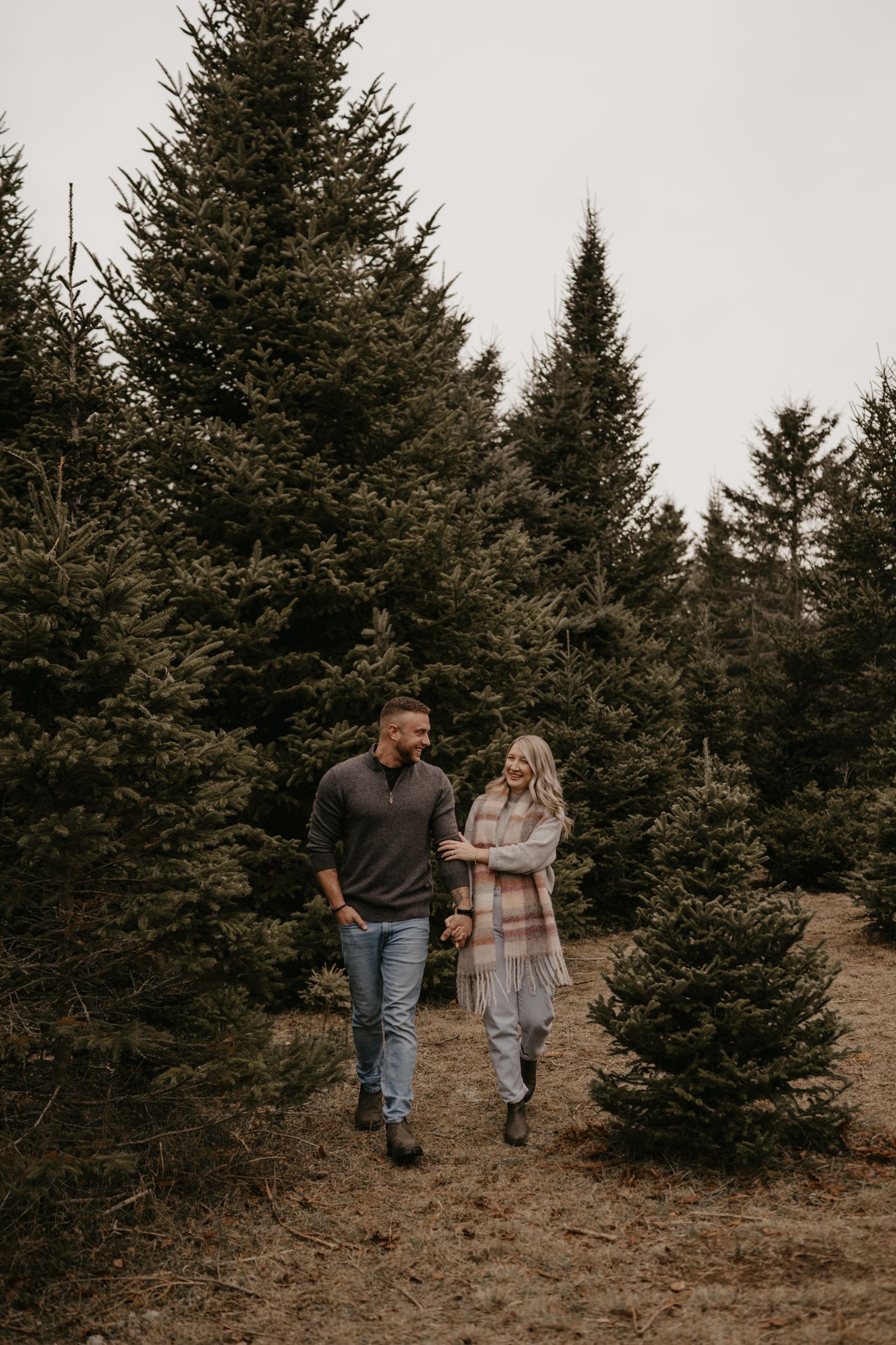 Couple walking hand in hand through a Christmas tree farm, wearing cozy fall outfits among tall evergreen trees.