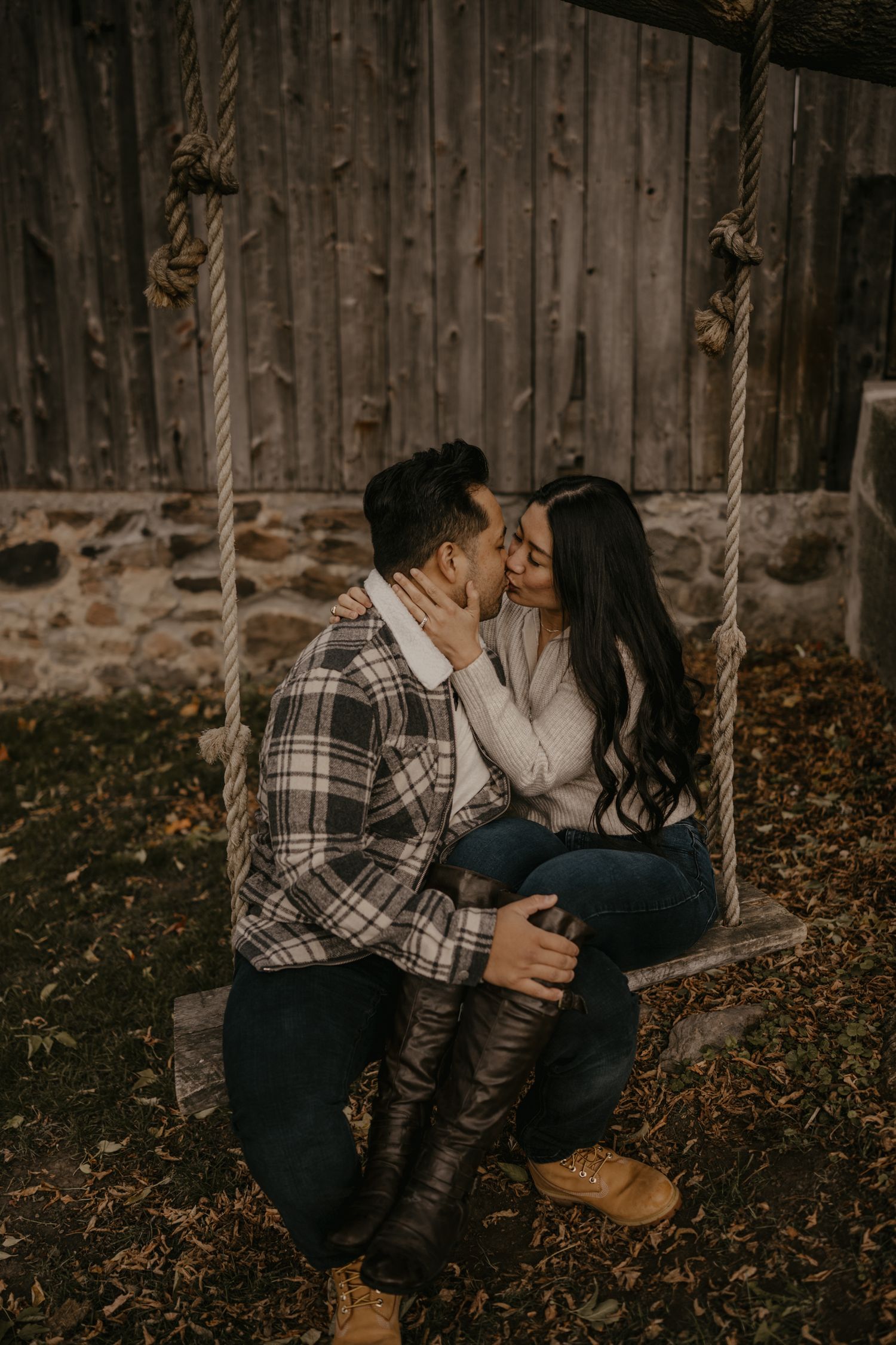 Couple shares a kiss on a rustic rope swing beside a stone barn in fall.