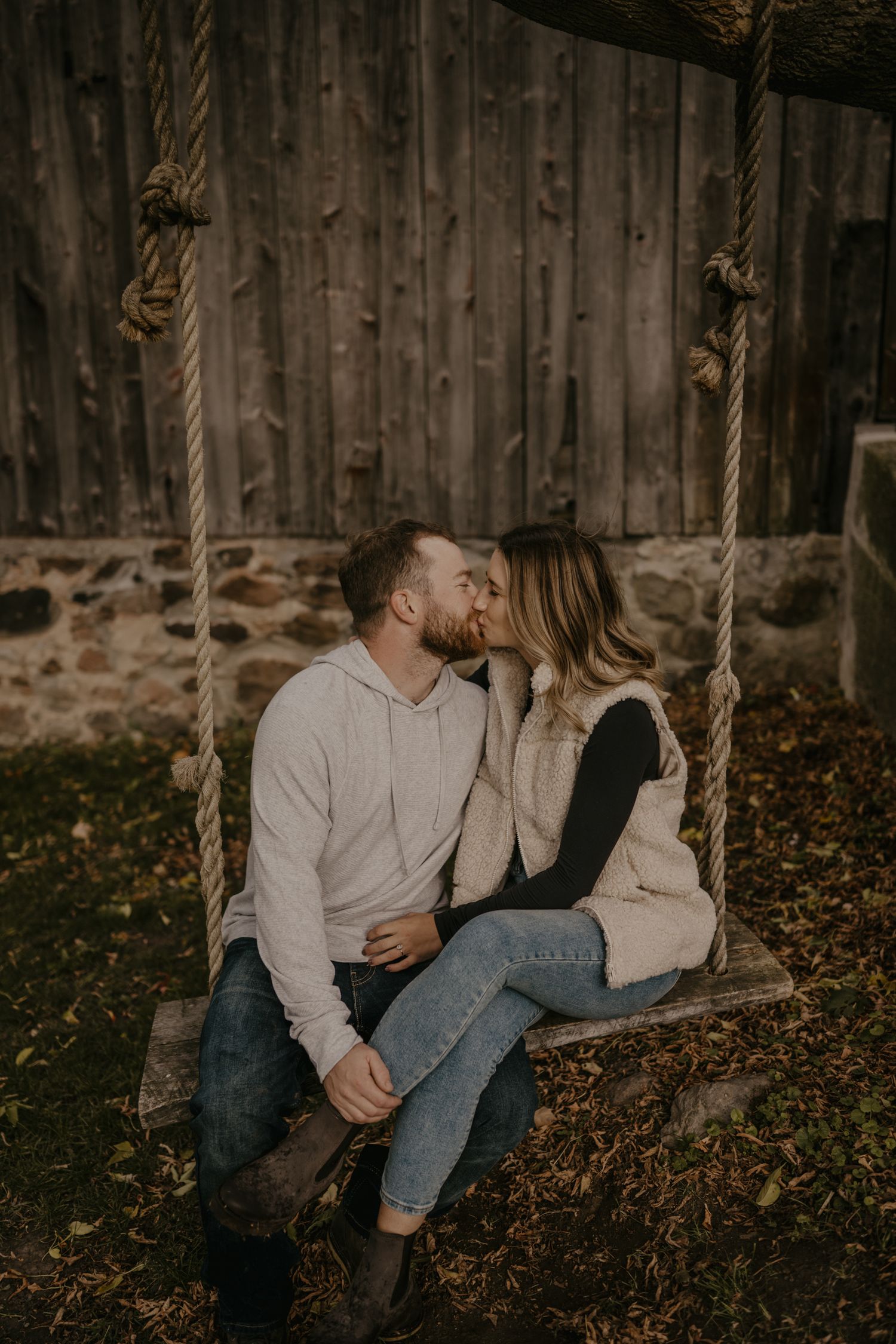 A couple shares a romantic kiss while sitting on a rustic rope swing outdoors in autumn.