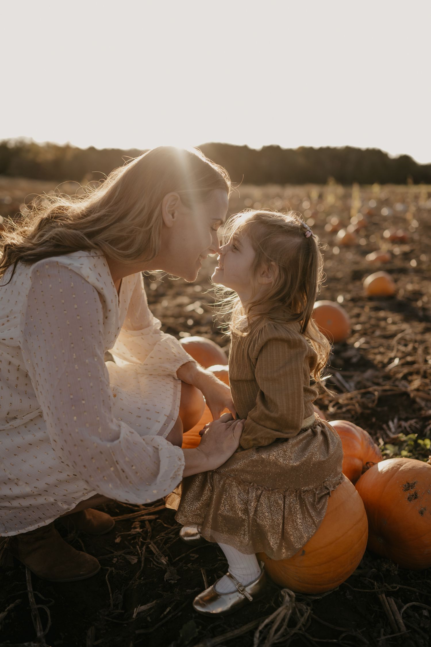 Mother and toddler daughter share a tender nose-to-nose moment in a sunlit pumpkin patch at golden hour.