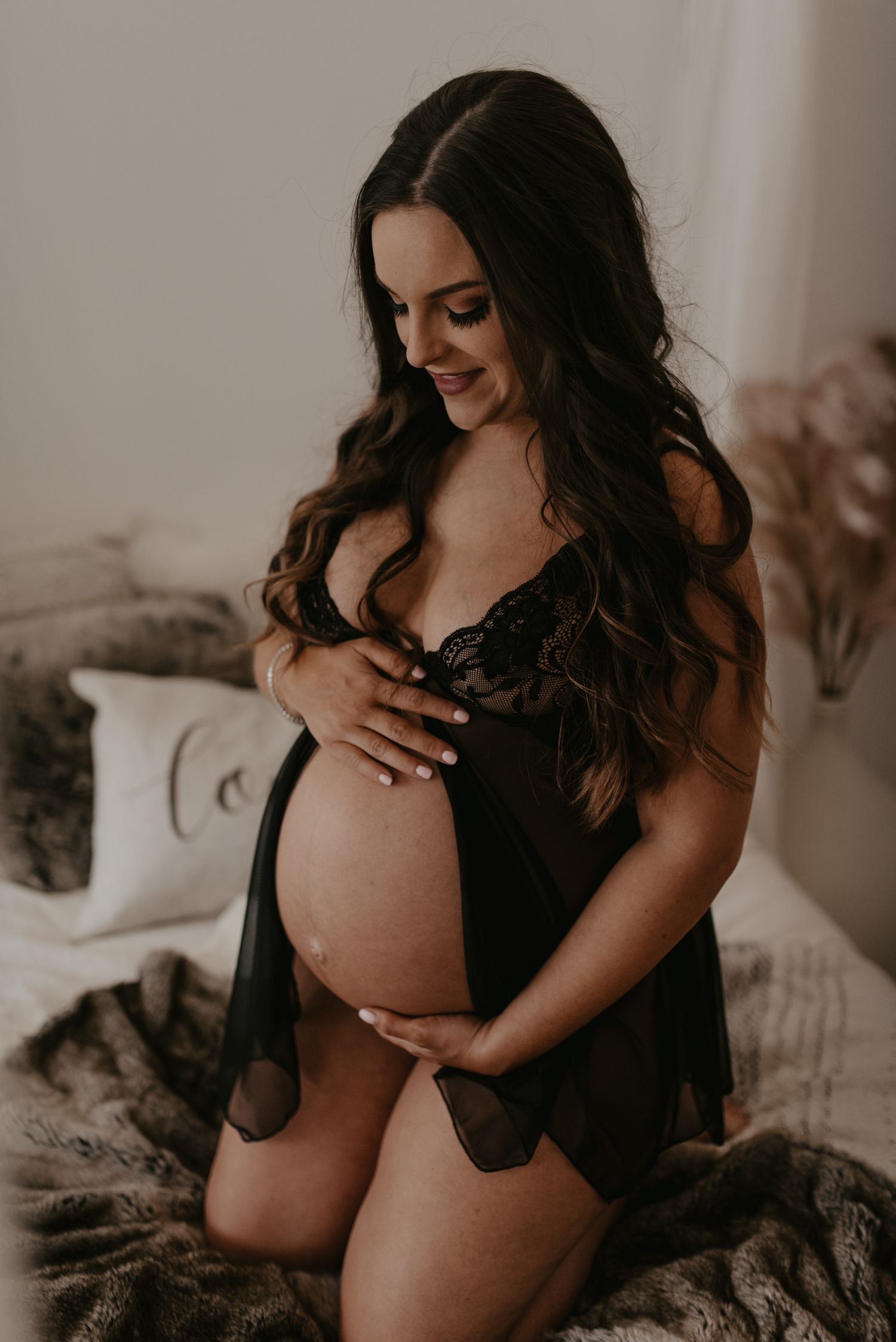 Smiling pregnant woman in black lace lingerie cradling her baby bump while kneeling on a bed.