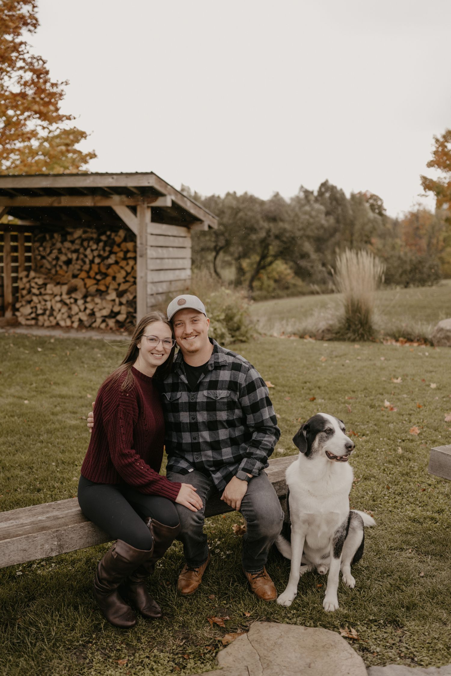 Couple smiling with black and white dog outdoors near rustic wood shed in autumn.