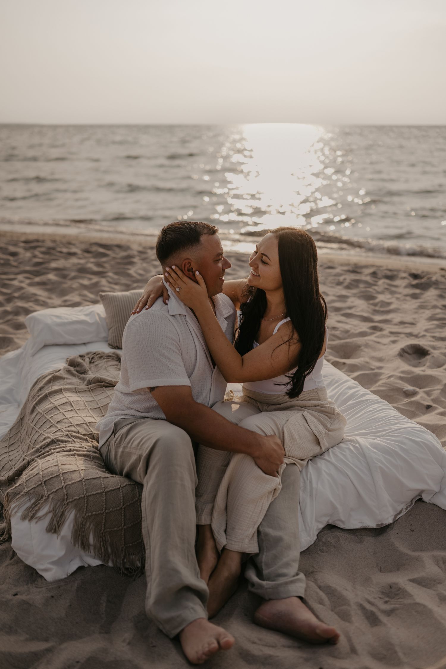 Romantic couple sitting on beach blanket at sunset, woman tenderly holding man's face.