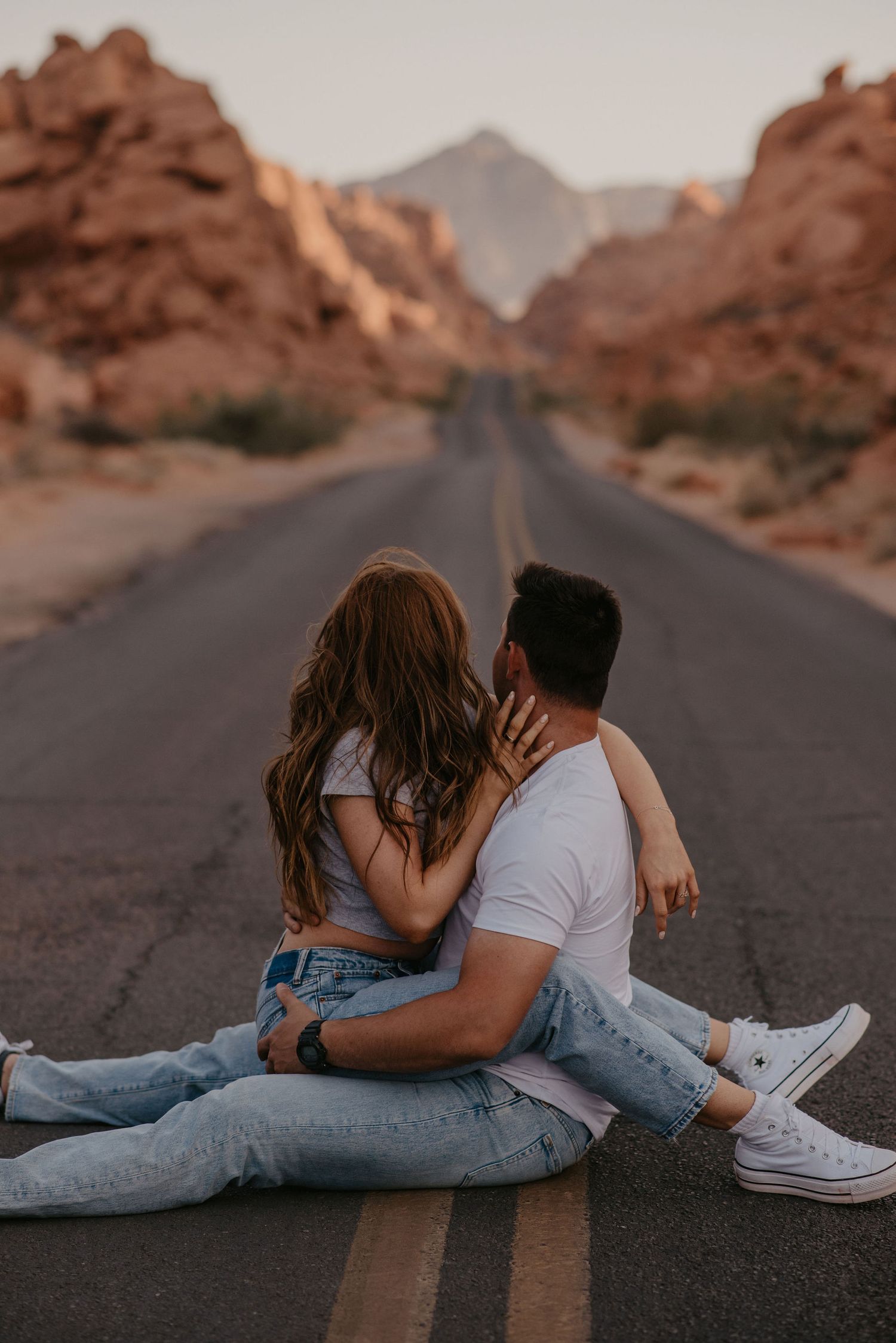 Couple sitting together on an empty desert highway surrounded by red rock canyon walls.