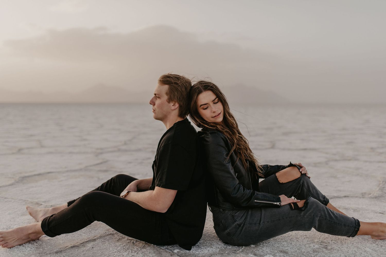 Couple sitting back to back on the Bonneville Salt Flats in matching dark outfits.