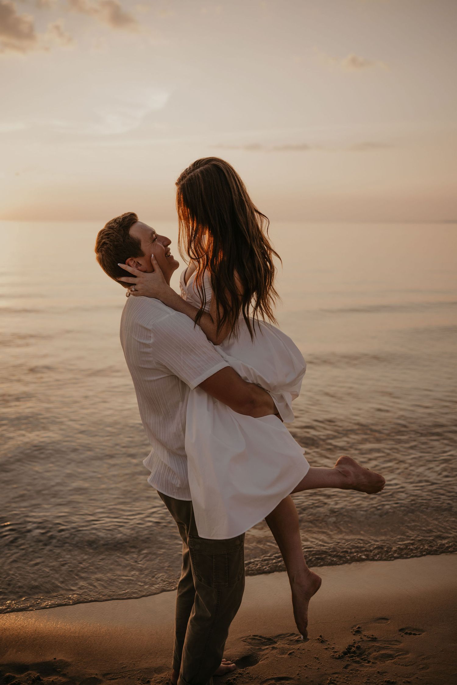 Couple sharing a romantic moment at sunset on the beach, man lifting woman in white dress by the ocean.