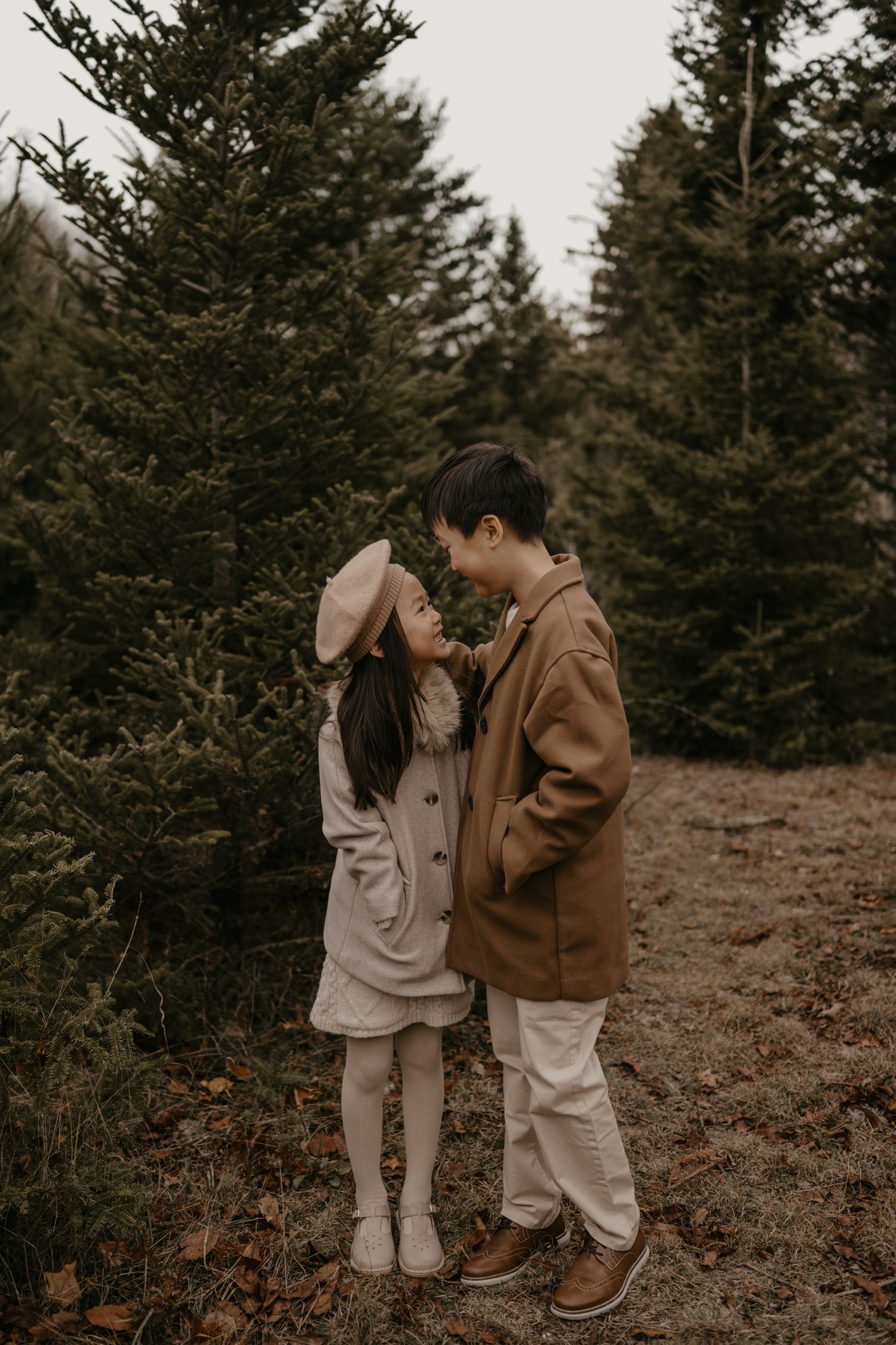 Two children in coordinating neutral coats share a sweet moment among evergreen trees outdoors.