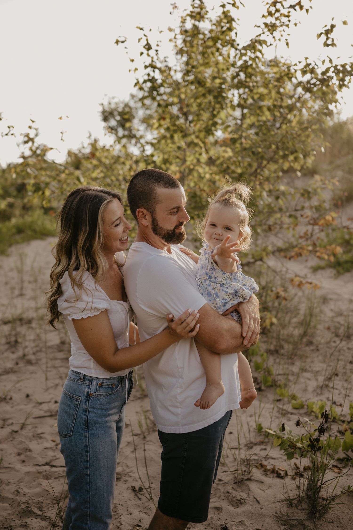 Young family of three standing together on sandy dunes with father holding baby girl at sunset.