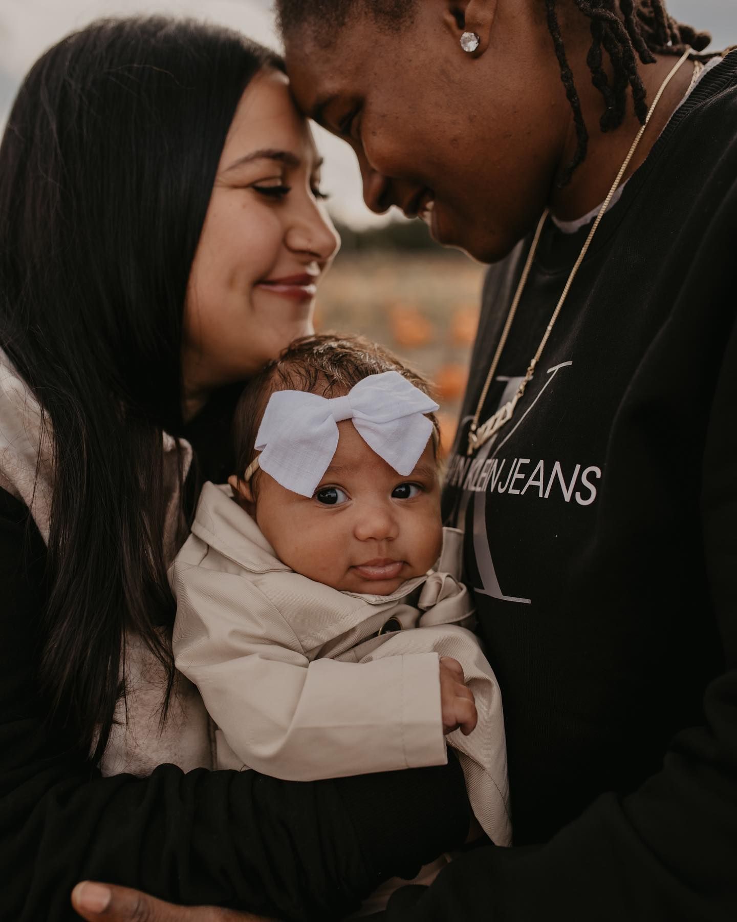 Young couple tenderly touching foreheads while holding their newborn baby with a white bow headband.