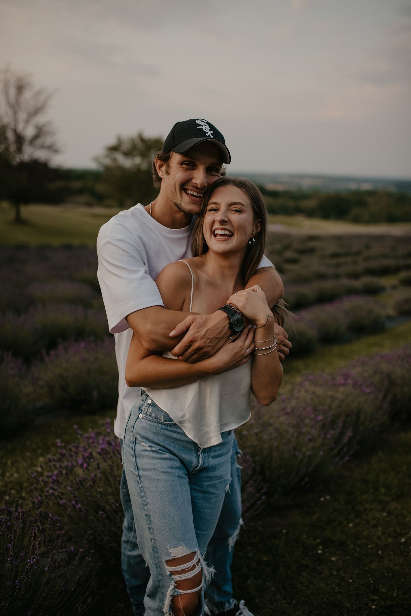 Happy couple embracing and laughing in a blooming lavender field at dusk in casual outfits.