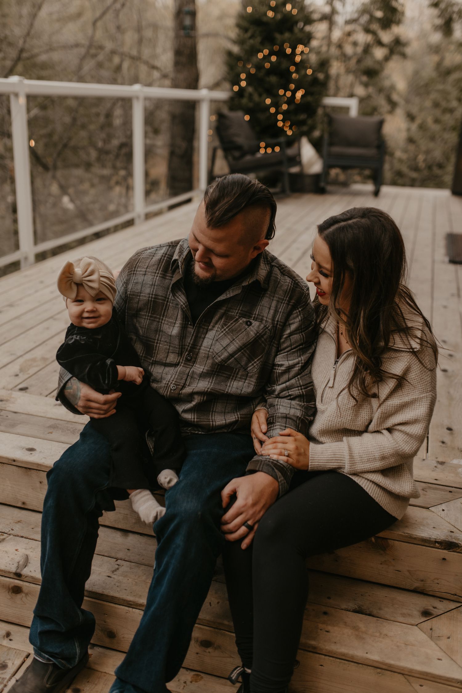 A young family sits on a wooden deck, parents smiling at their toddler with a Christmas tree glowing in the background.