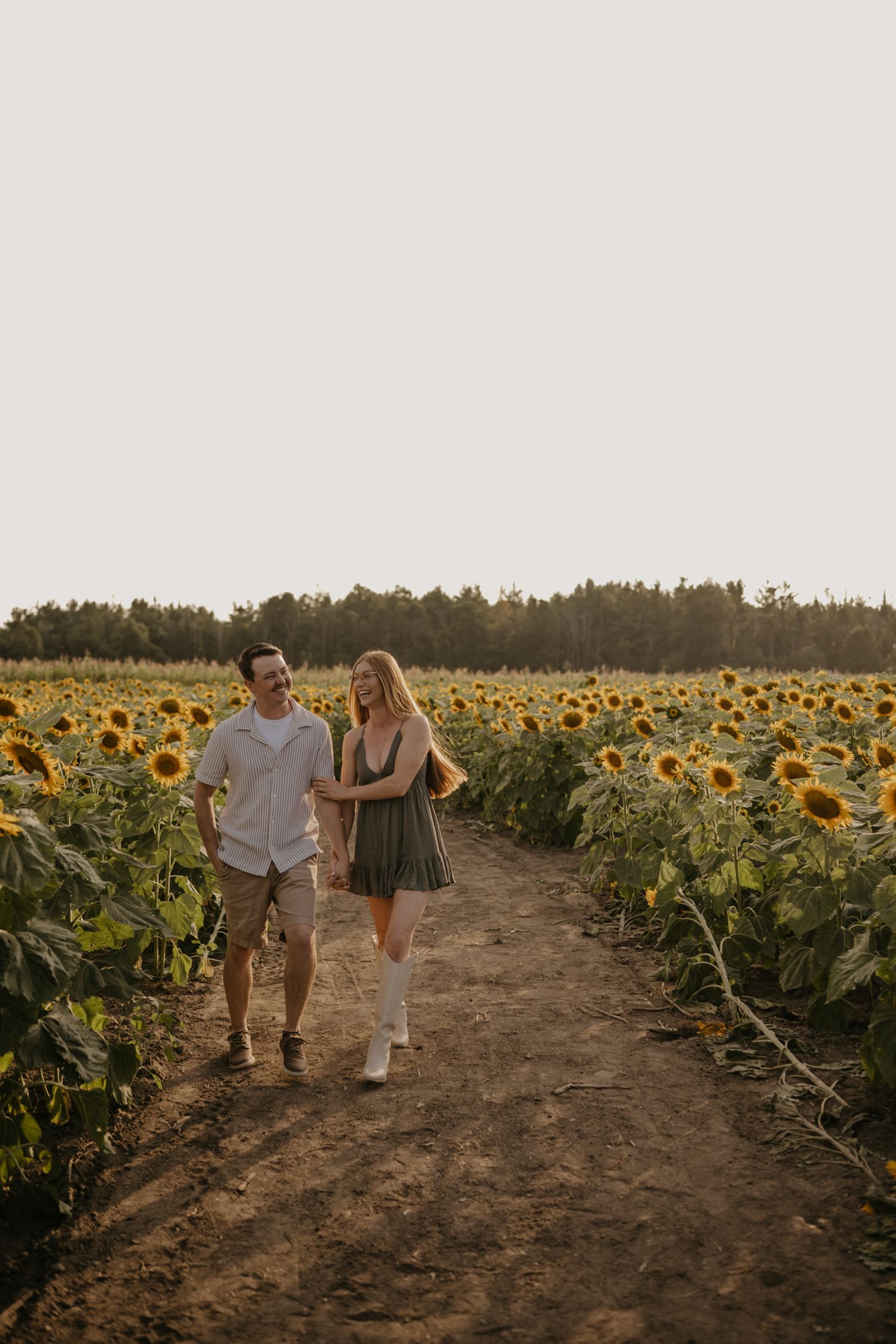 Smiling couple walking hand in hand along a dirt path through a vast sunflower field at sunset.