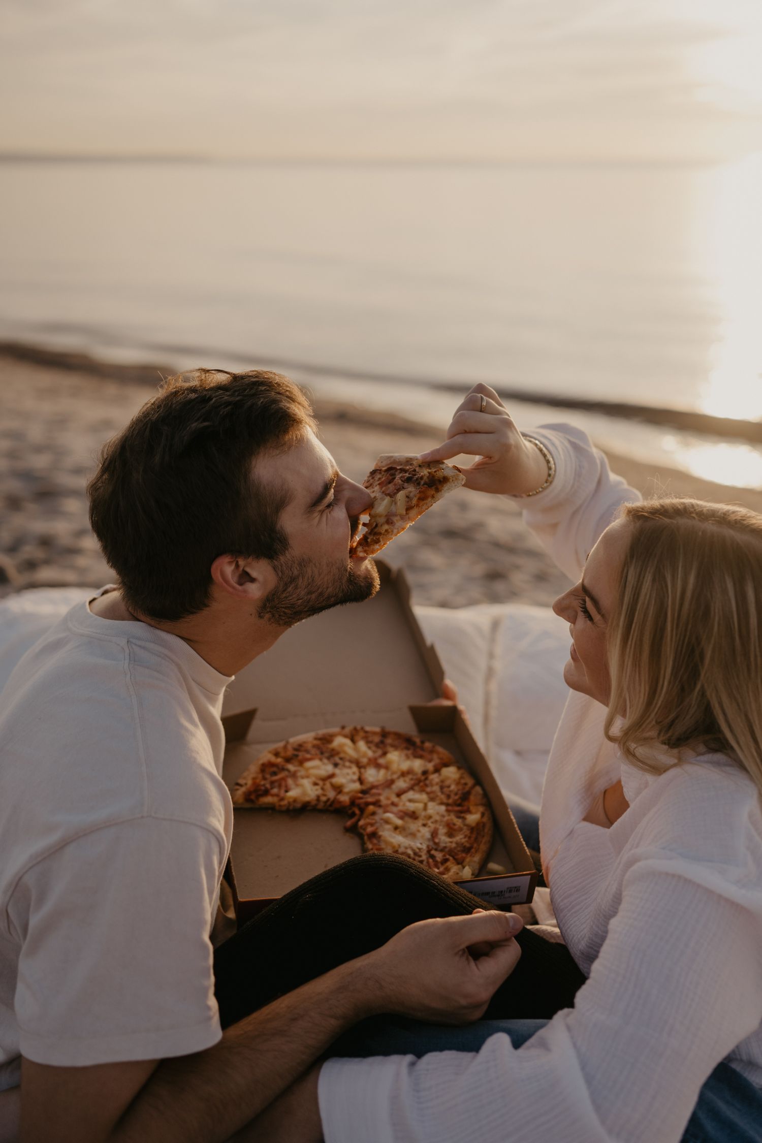 Couple sharing pizza on beach blanket at sunset, woman feeding man a slice romantically.
