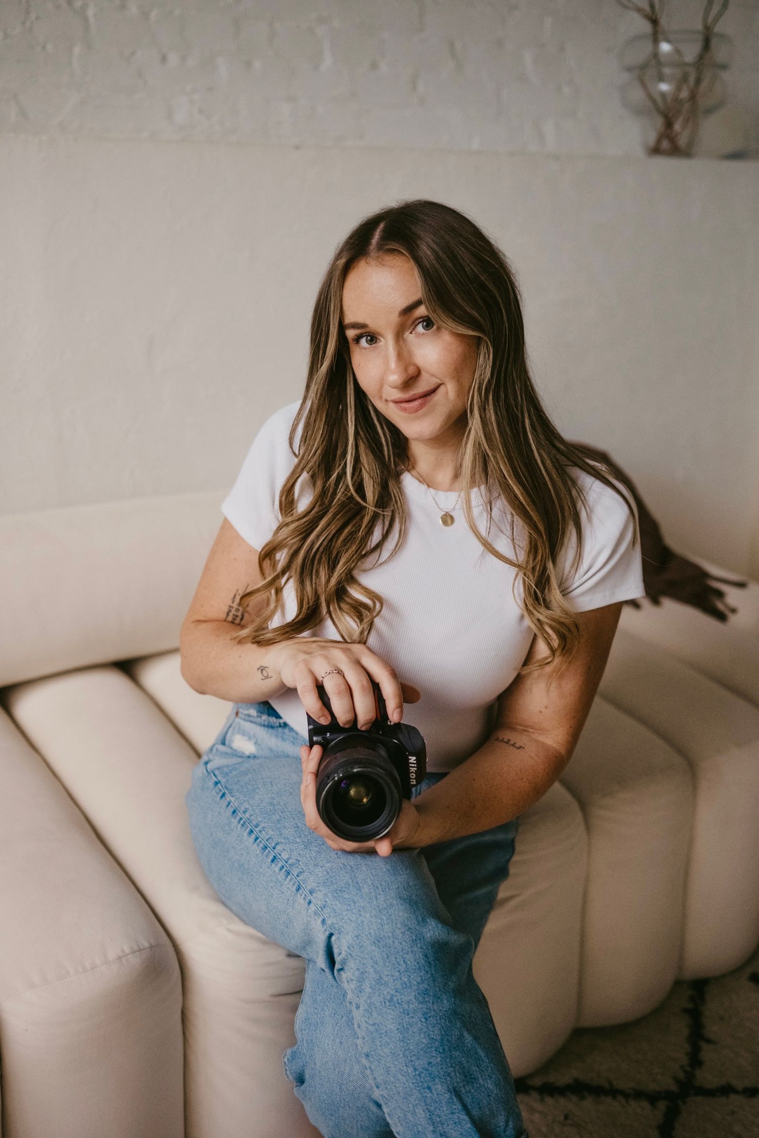 Female photographer sitting on cream sofa holding DSLR camera in bright white studio setting.