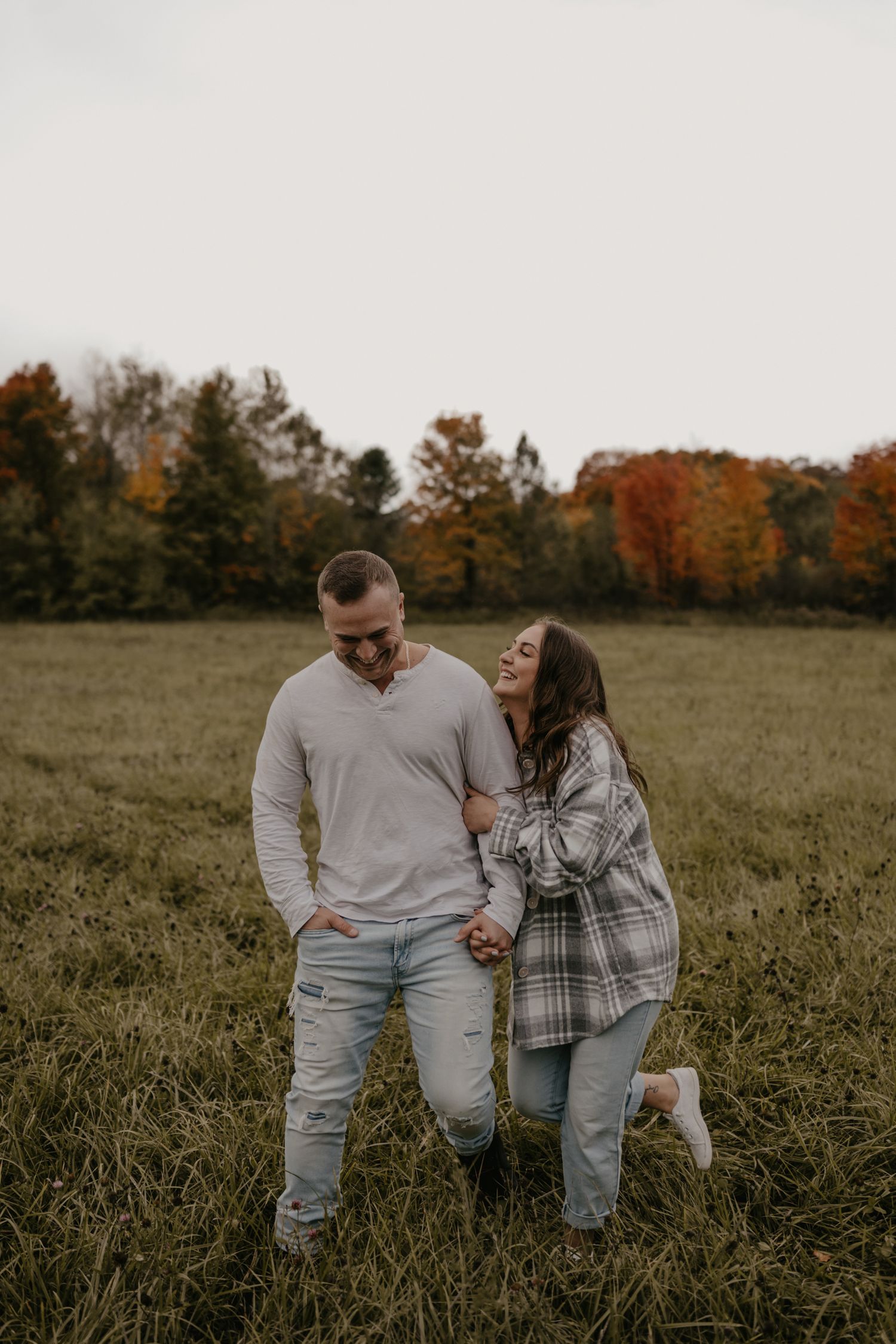 Couple laughing together in a green field with colorful autumn trees, man in gray shirt and woman in plaid jacket.