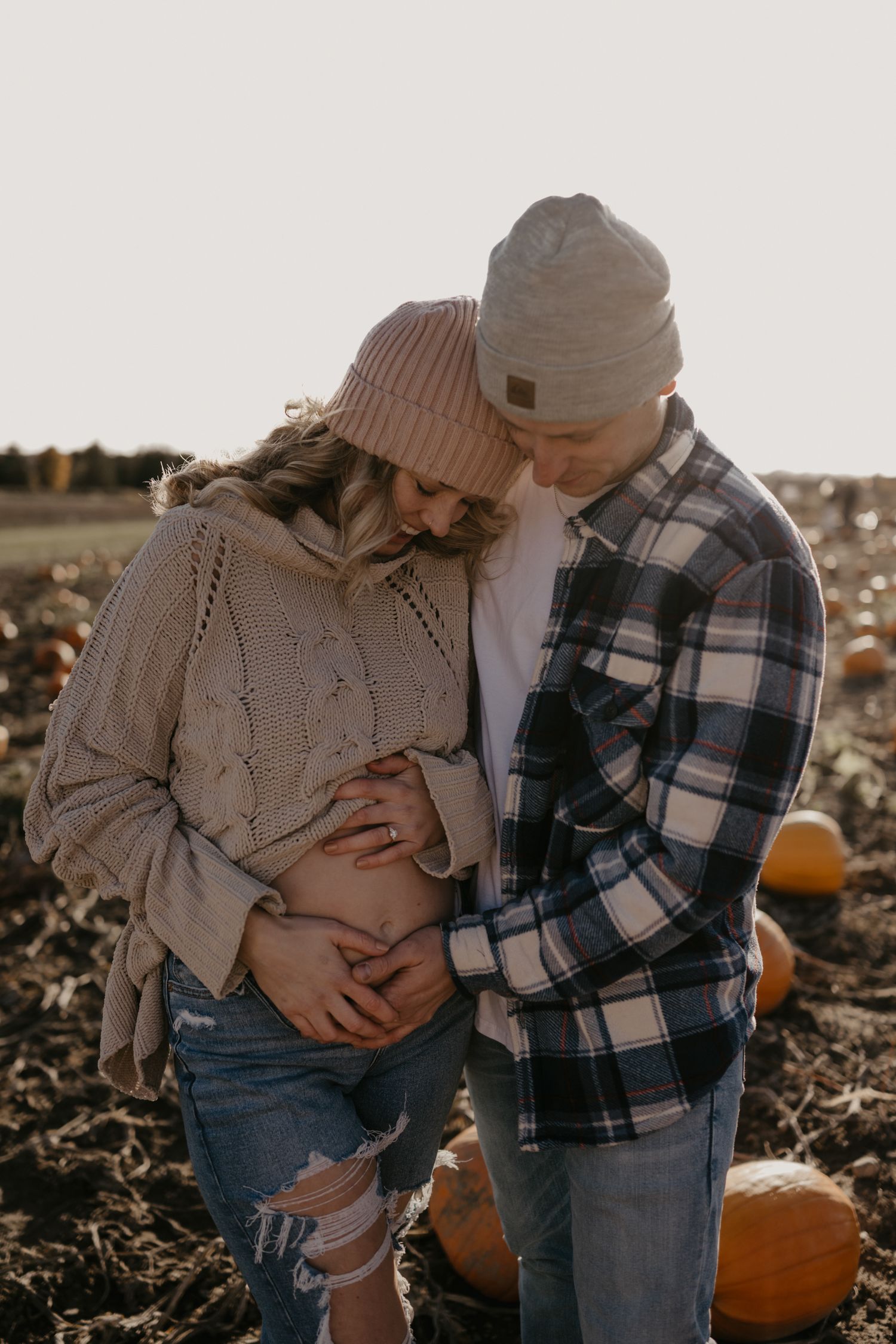 Couple in beanies and cozy sweaters embracing in a pumpkin patch, cradling a baby bump during a fall maternity photoshoot.
