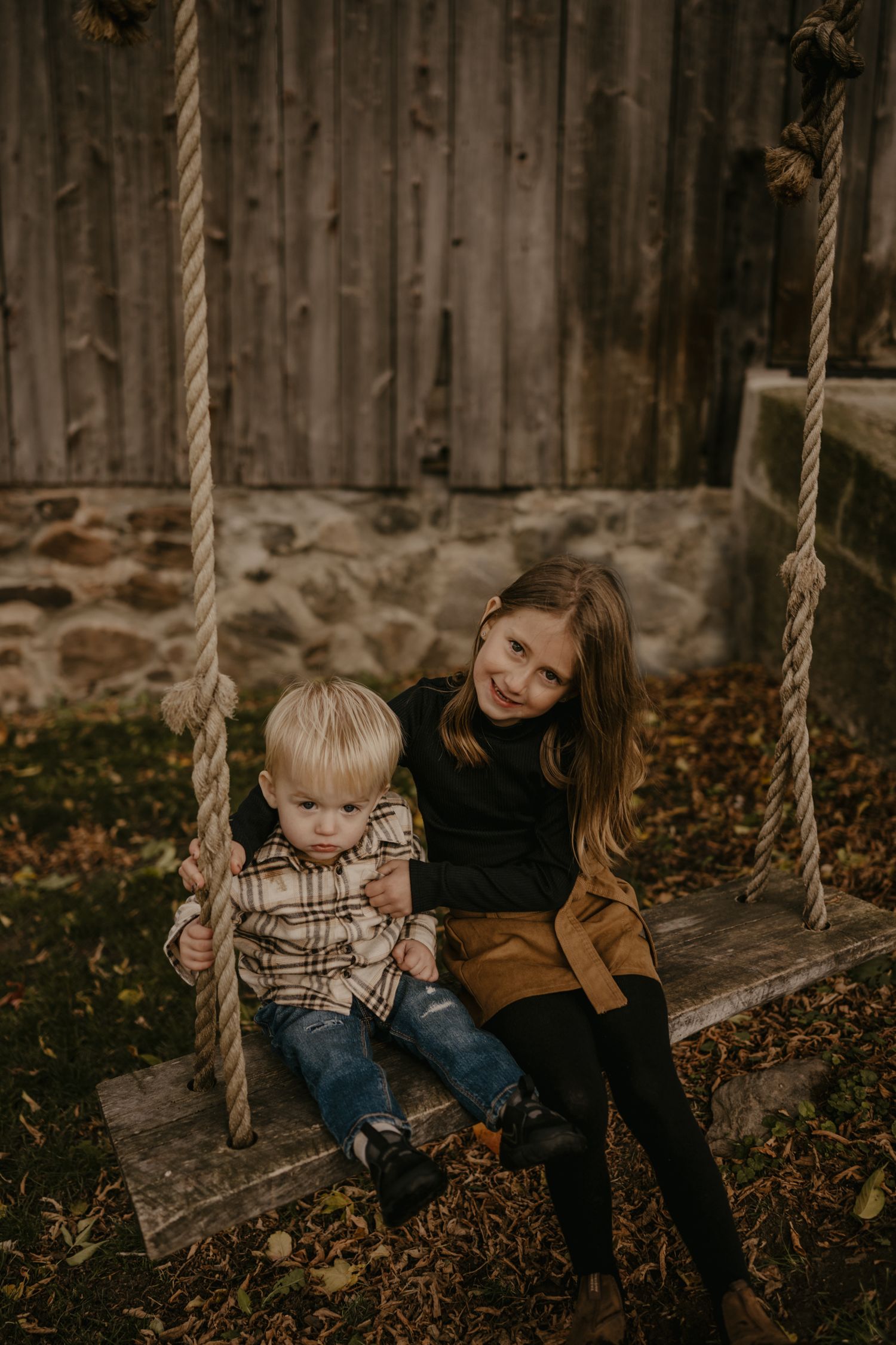 Two young children sitting together on a wooden rope swing in fall setting.