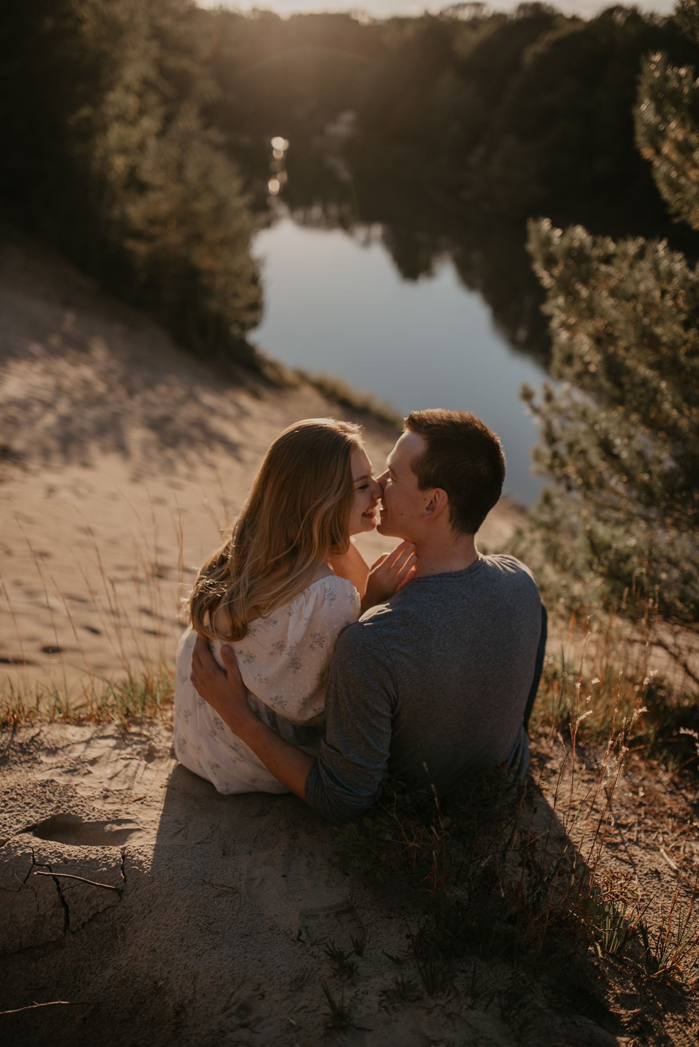 Couple sharing a tender almost-kiss while sitting on sandy dunes overlooking a peaceful river at sunset.