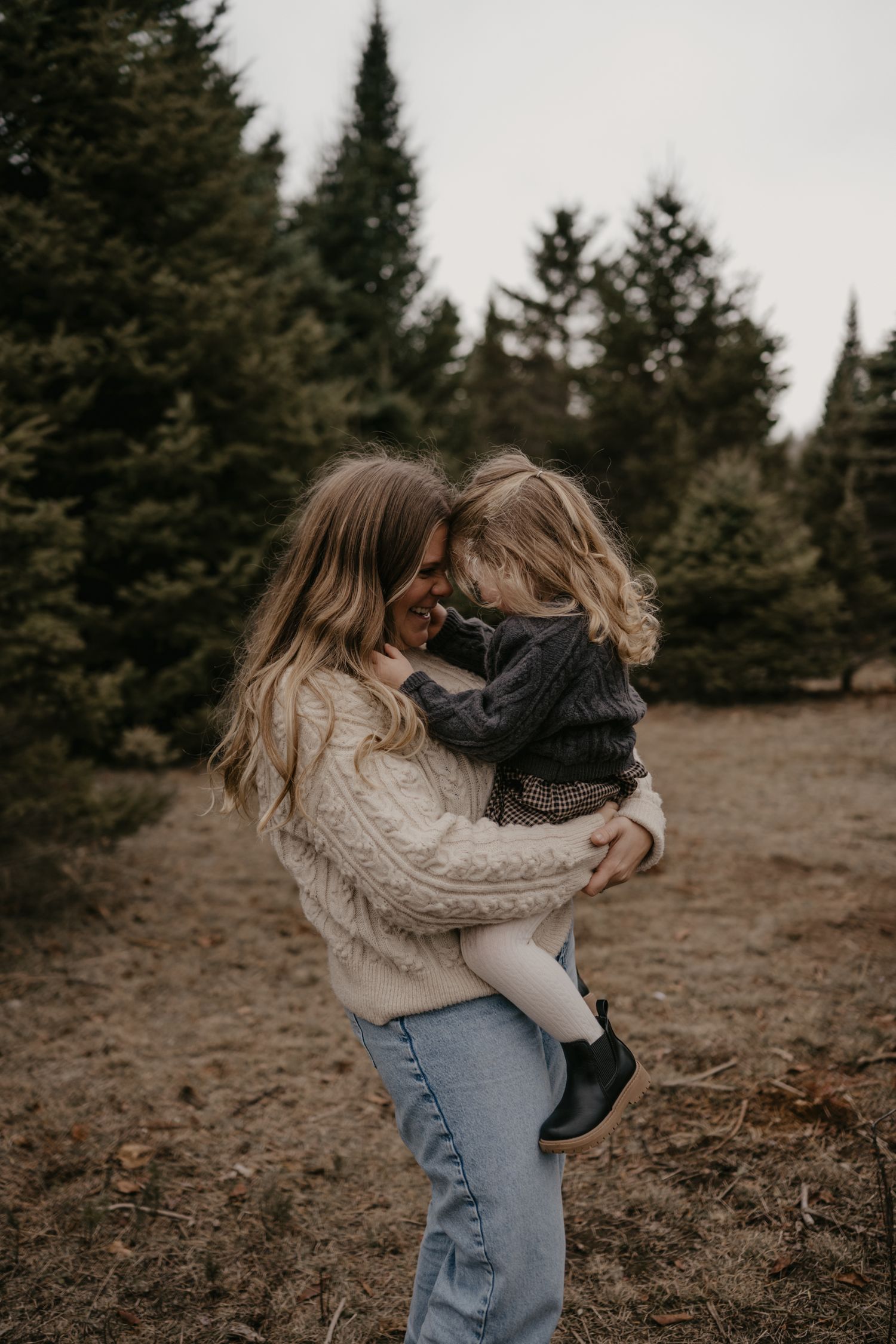 A mother holding her young child in a warm embrace outdoors near evergreen trees in a cozy knit sweater.