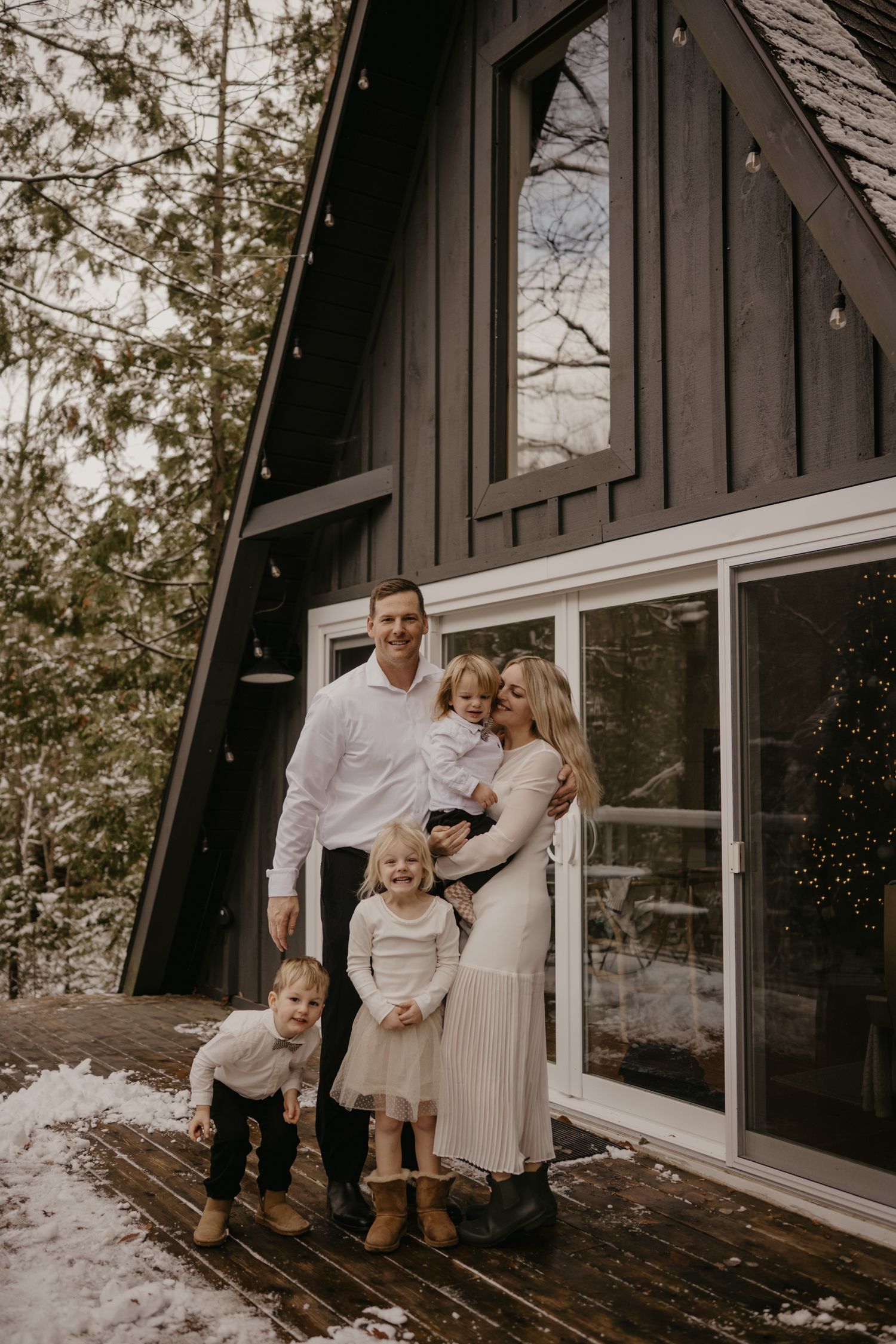 Happy family of five posing outside a dark A-frame cabin on a snowy winter day, dressed in coordinated outfits.